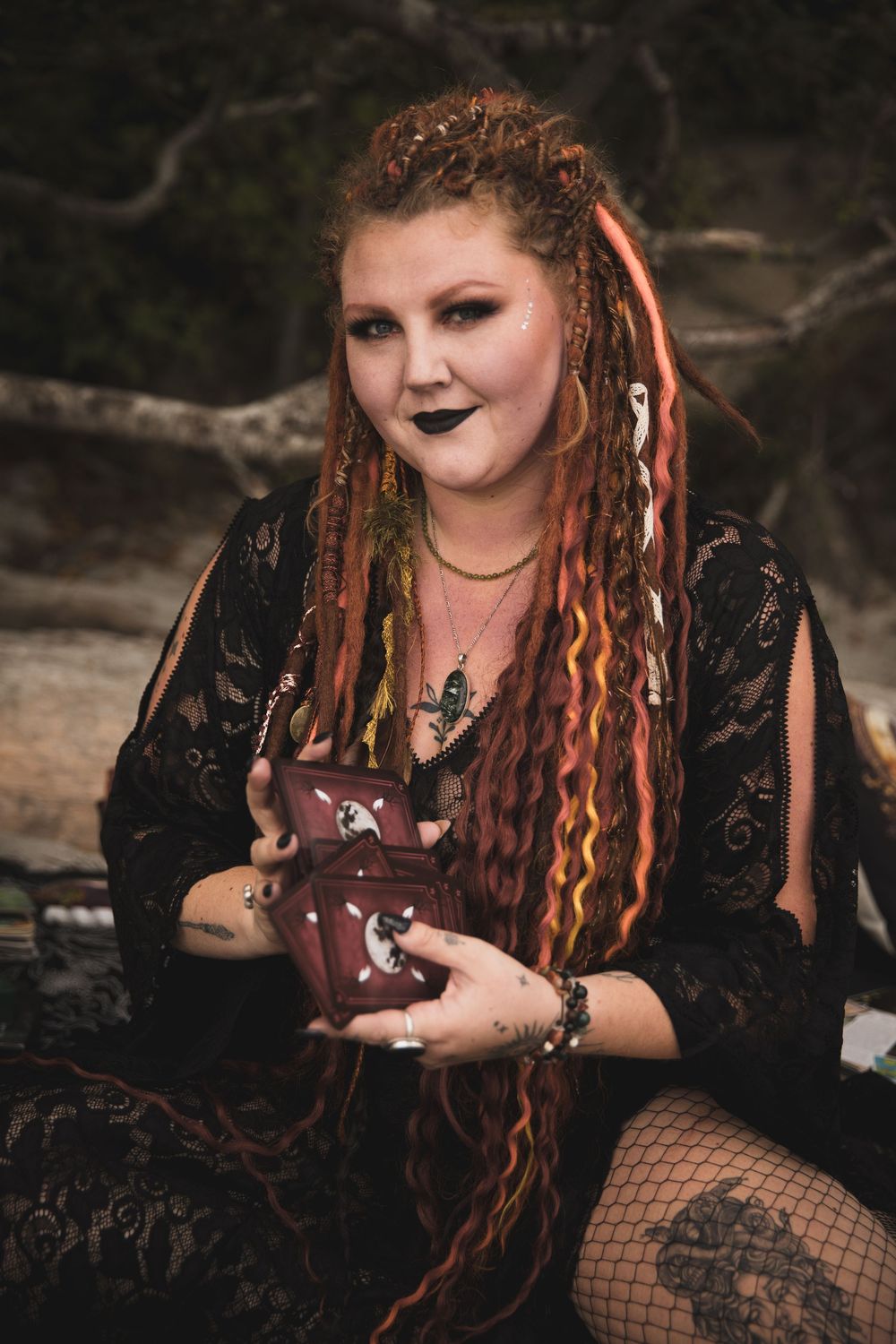 Gothic-styled person with dreadlocks wearing black lace dress and fishnet stockings holds small object in outdoor setting.