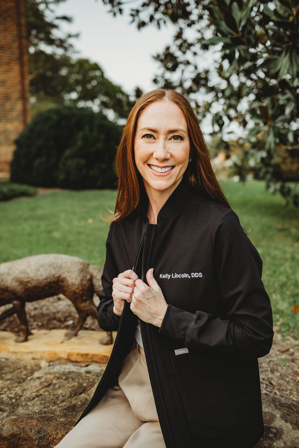 A person in a black coat sits outdoors on a sunny day, smiling warmly against a natural backdrop.