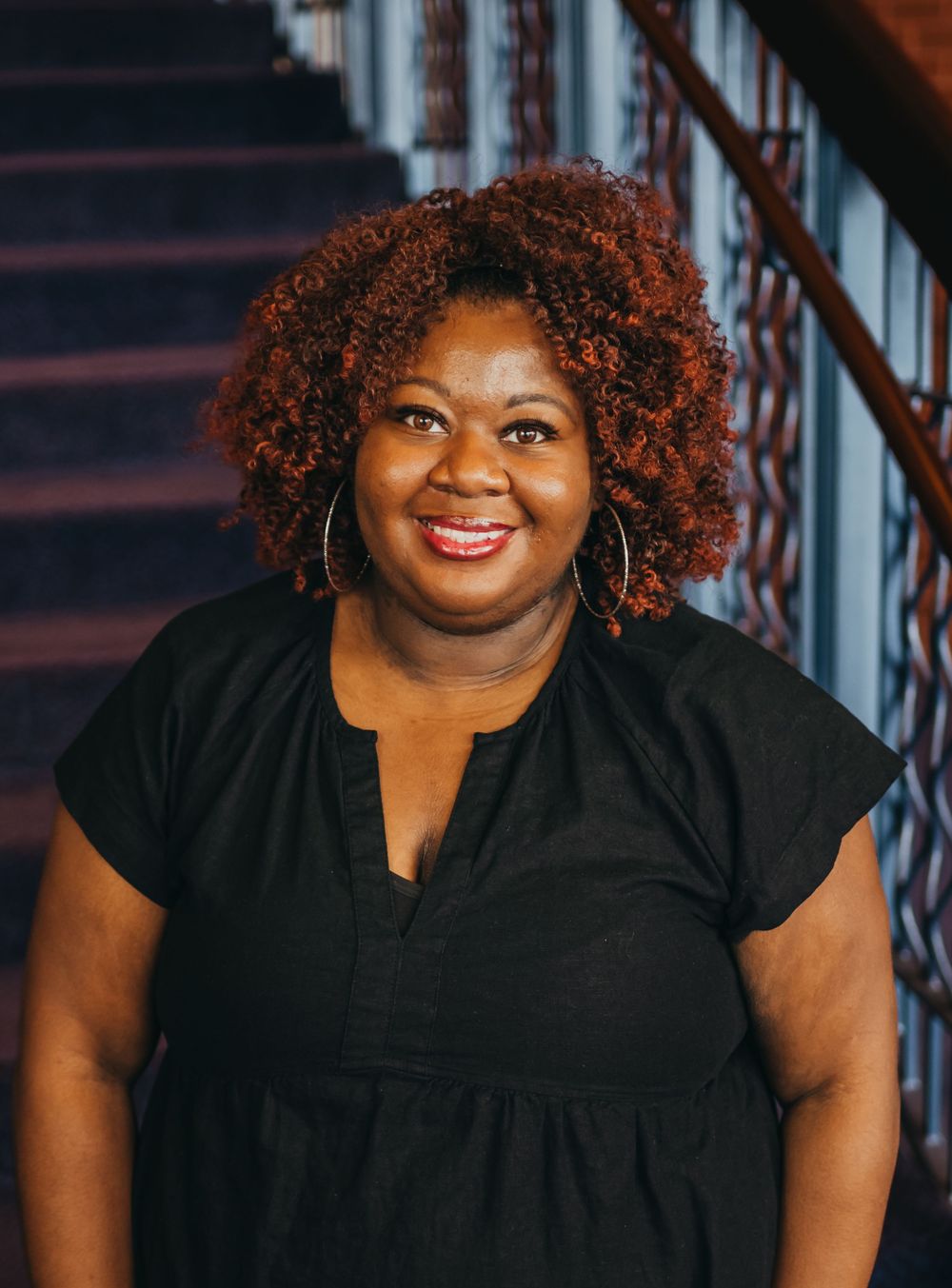 A smiling individual with vibrant red curly hair wearing a black top poses near a staircase.