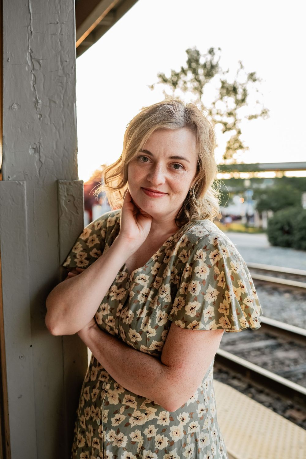 Person in a floral print dress poses thoughtfully near a train platform at sunset.