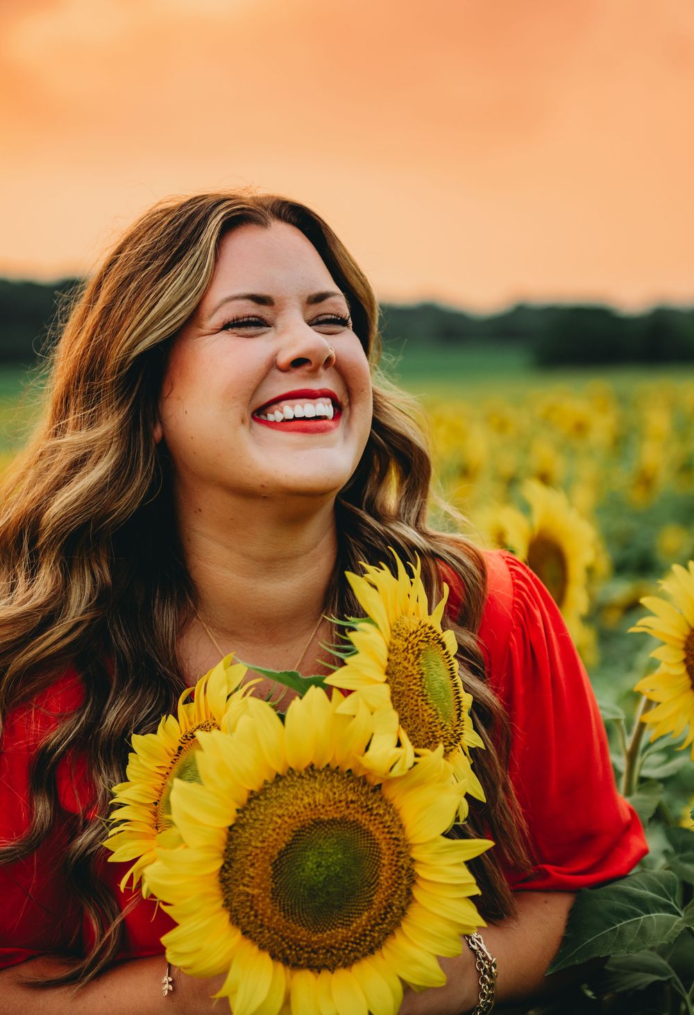 A joyful smile radiates in a sunflower field at sunset, with warm light and a vibrant red shirt against golden blooms.