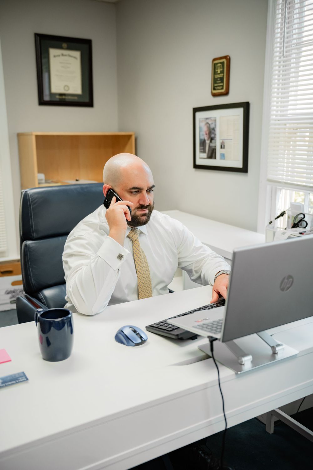 Bald businessman in white shirt and tie talking on phone while working on HP laptop at office desk.