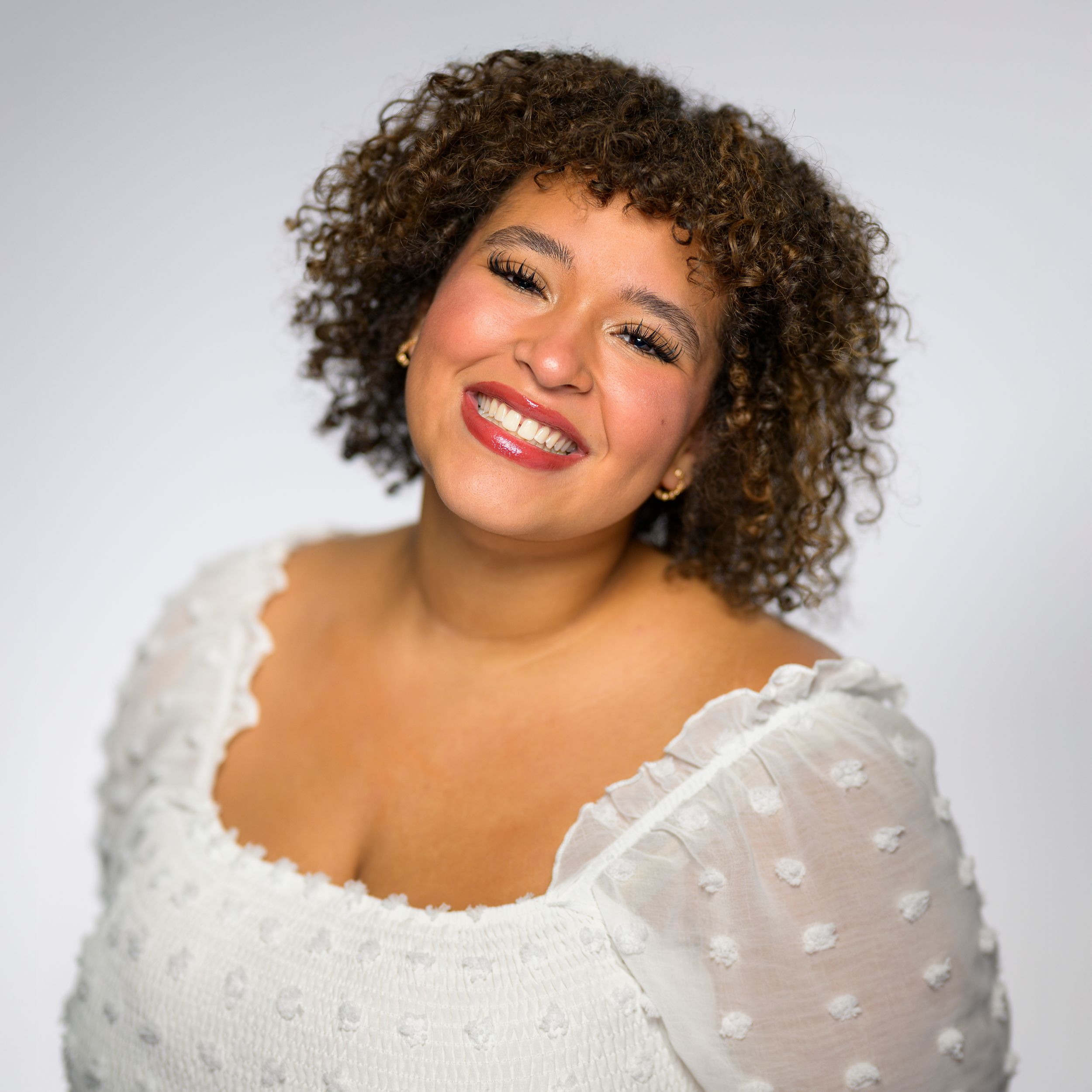 Bright studio portrait of a smiling young woman with curly hair in a white top.
