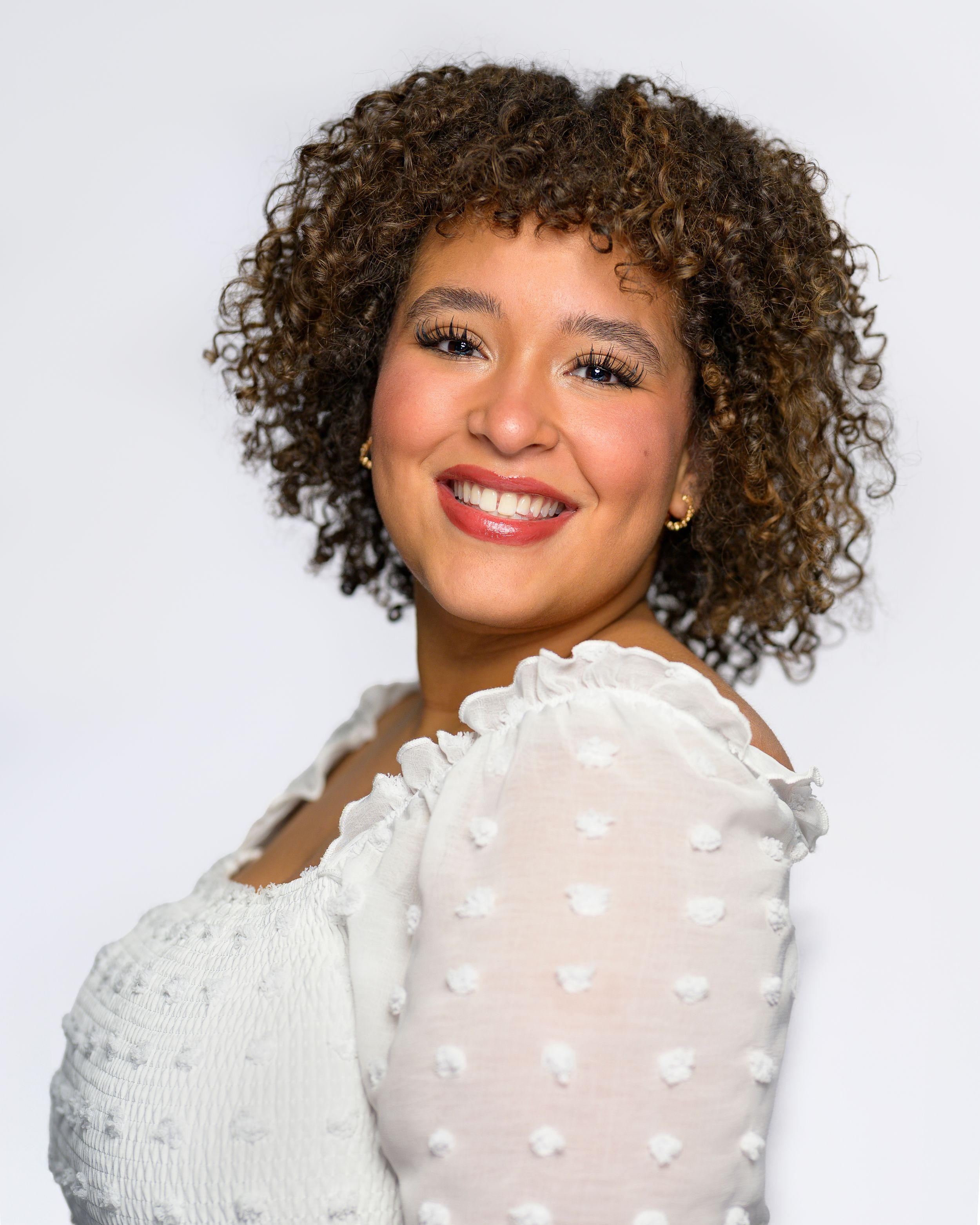 Side-angle studio portrait of a smiling young woman with curly hair in white top.