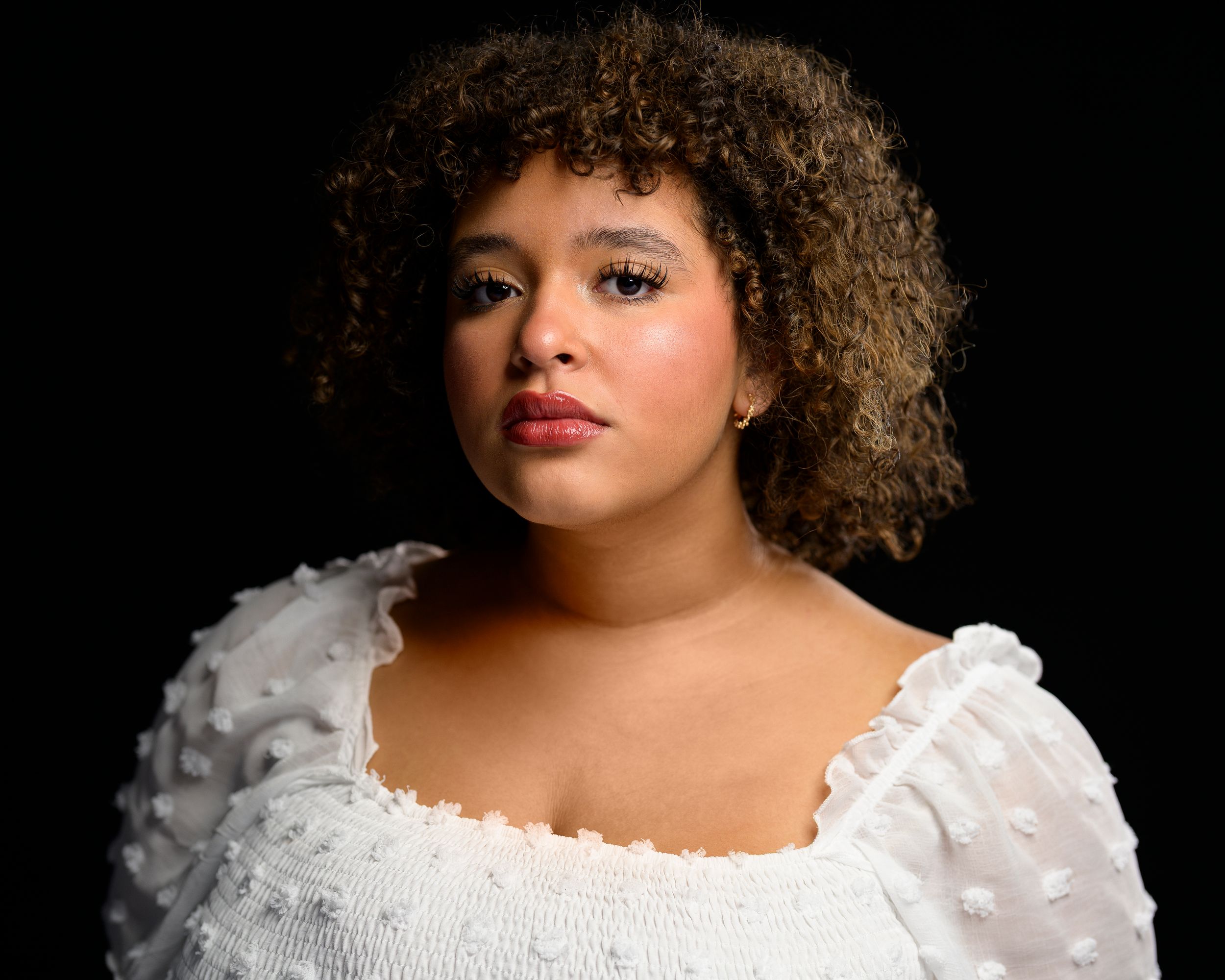 Young woman with curly hair wearing a white ruffled top poses against a dark background.