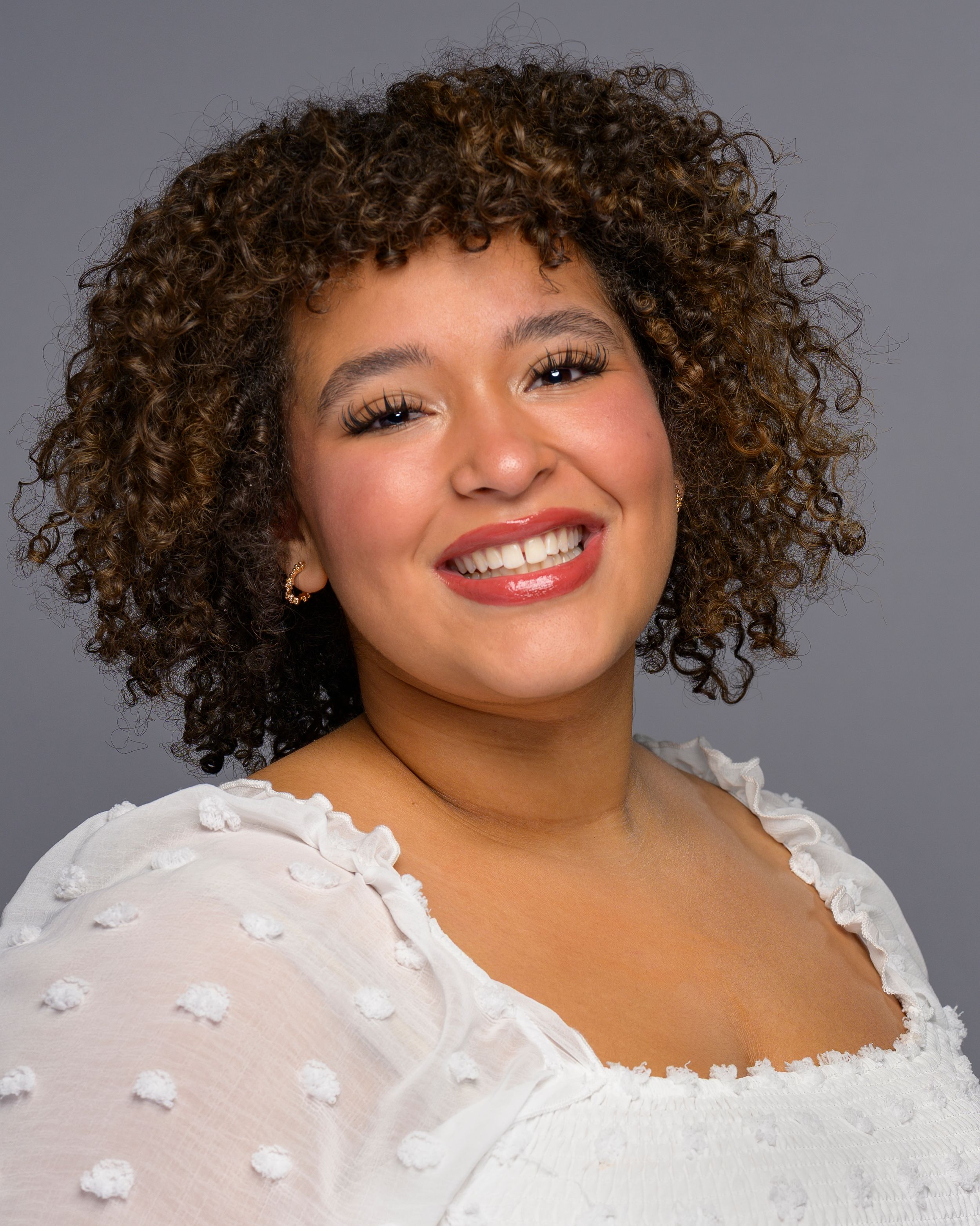 Portrait of a smiling young woman with curly hair wearing a white textured top.