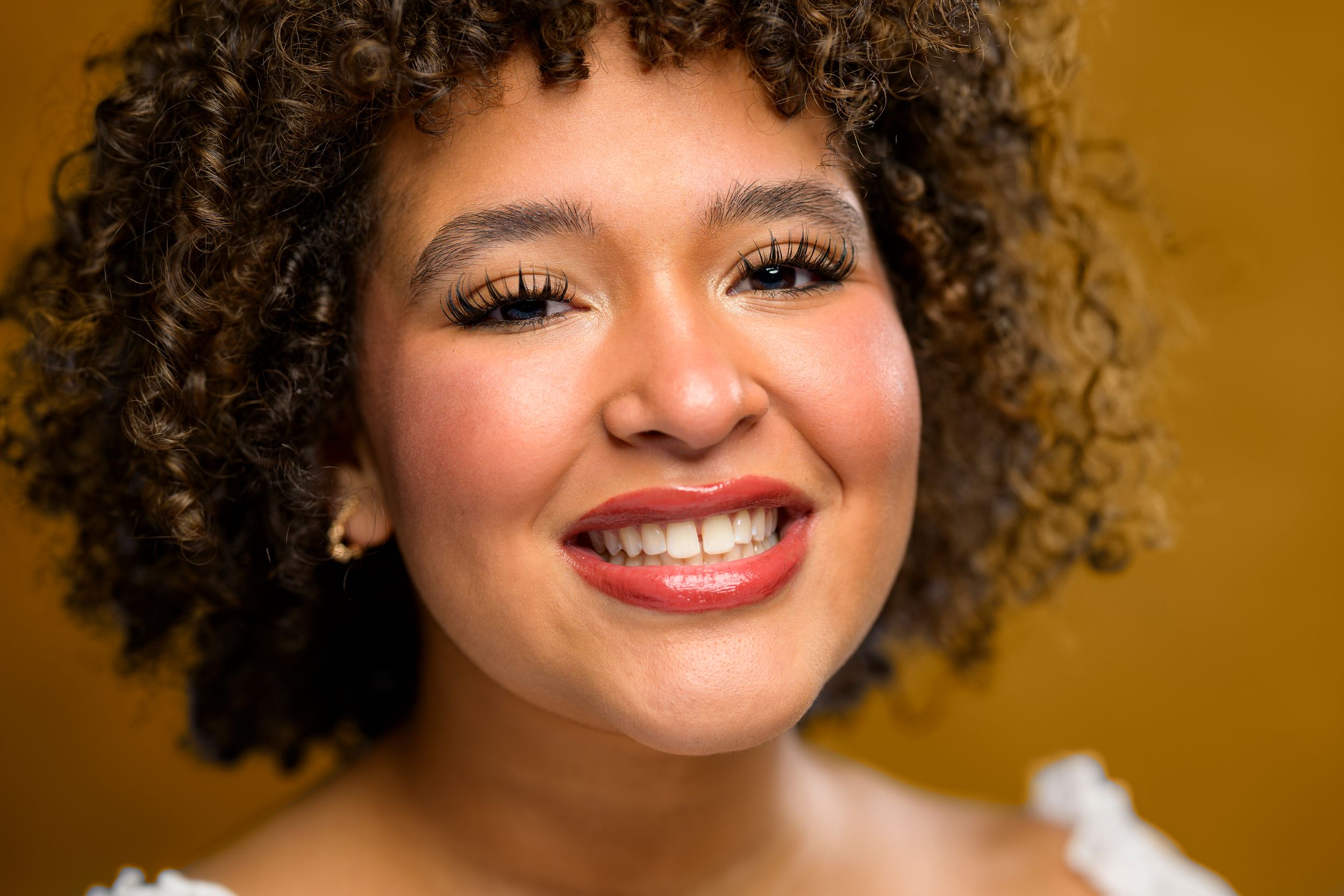Smiling young woman with curly hair and glittery eye makeup against a golden background.
