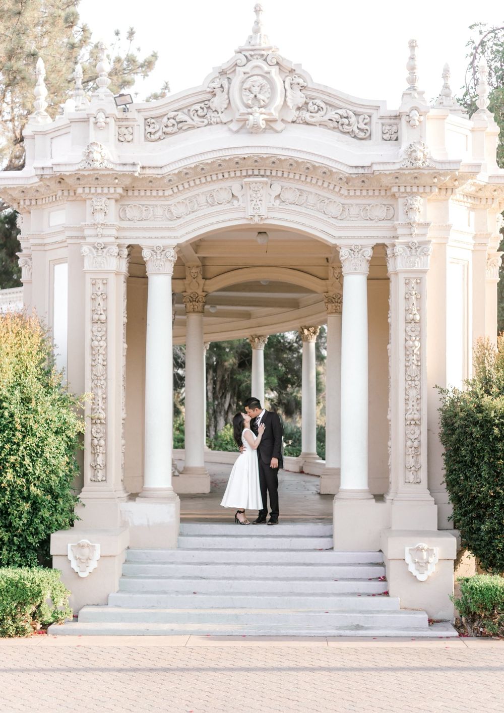 Couple sharing a romantic kiss under an ornate white baroque pavilion with columns and steps at Balboa Park.