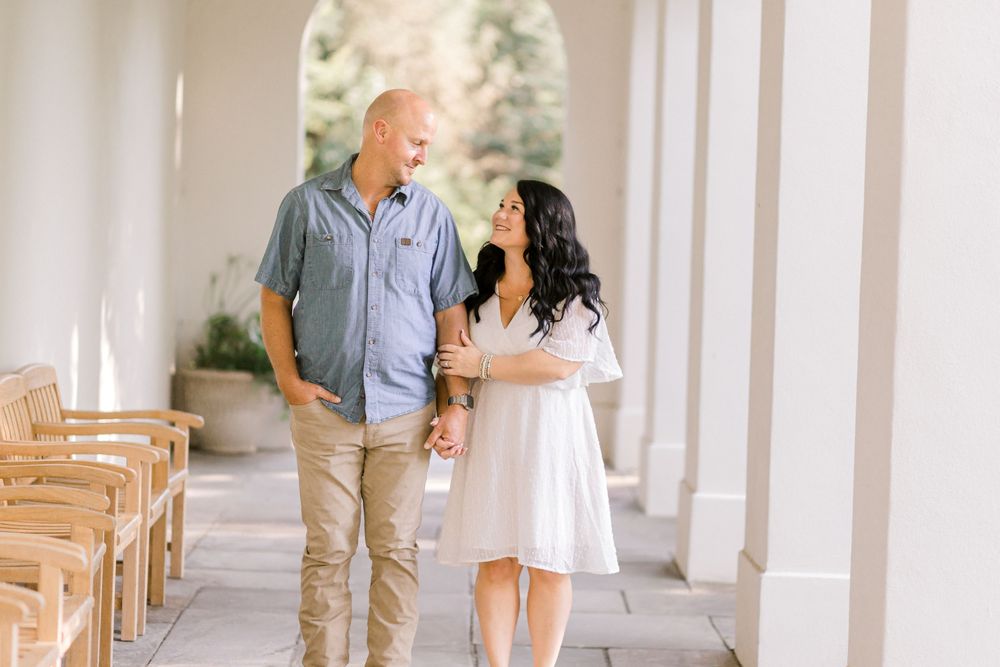 Couple holding hands and smiling while walking through elegant white columned outdoor corridor.