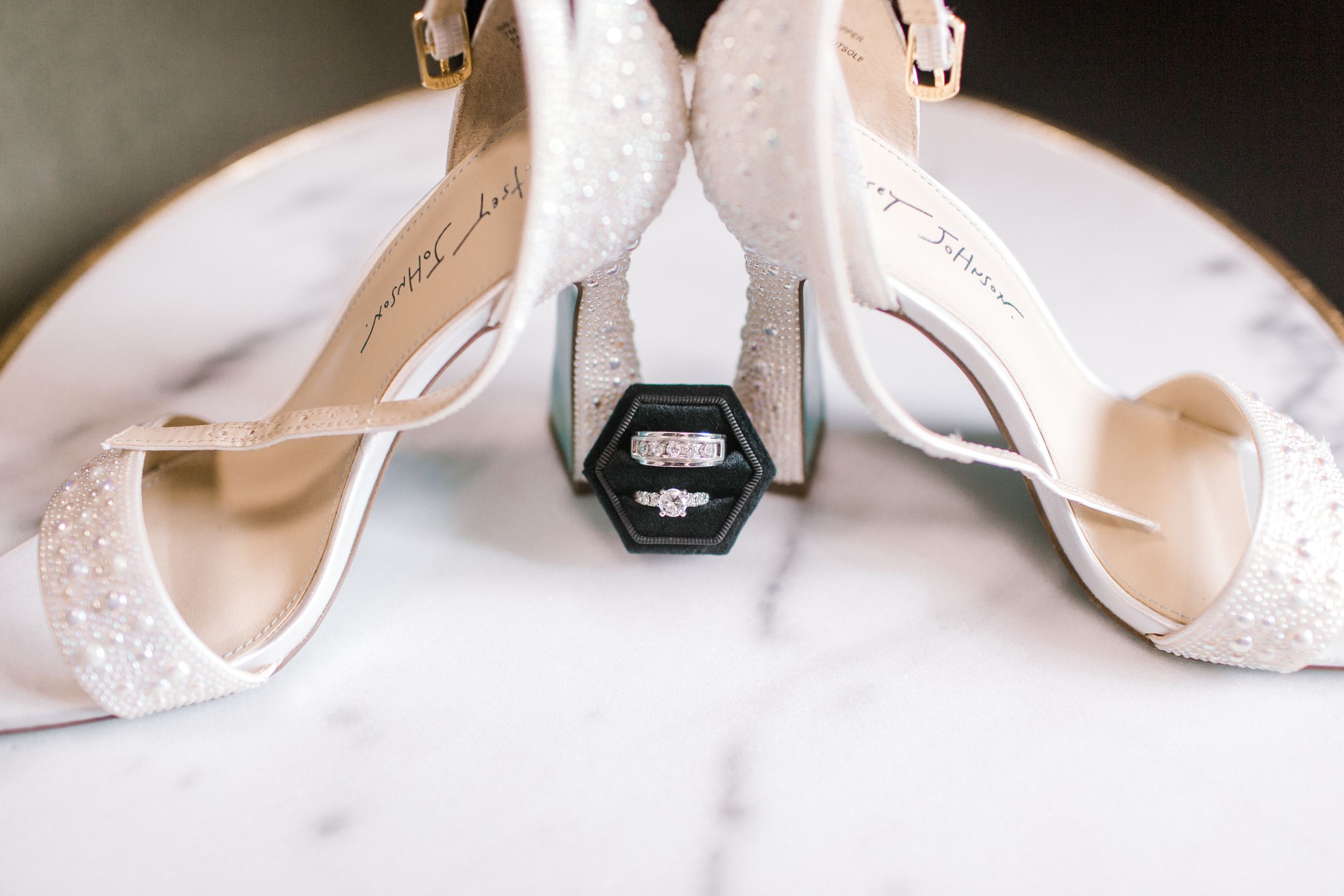 White bridal heels with pearl details framing a hexagonal ring box with engagement ring on marble surface.