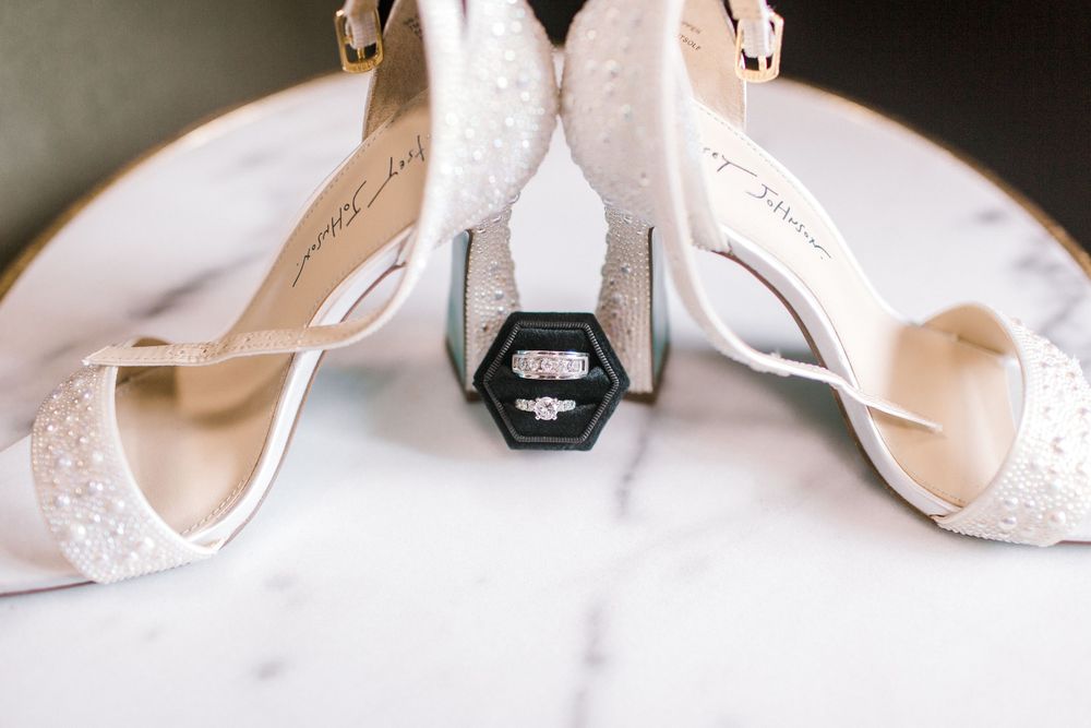 White bridal heels with pearl details framing a hexagonal ring box with engagement ring on marble surface.