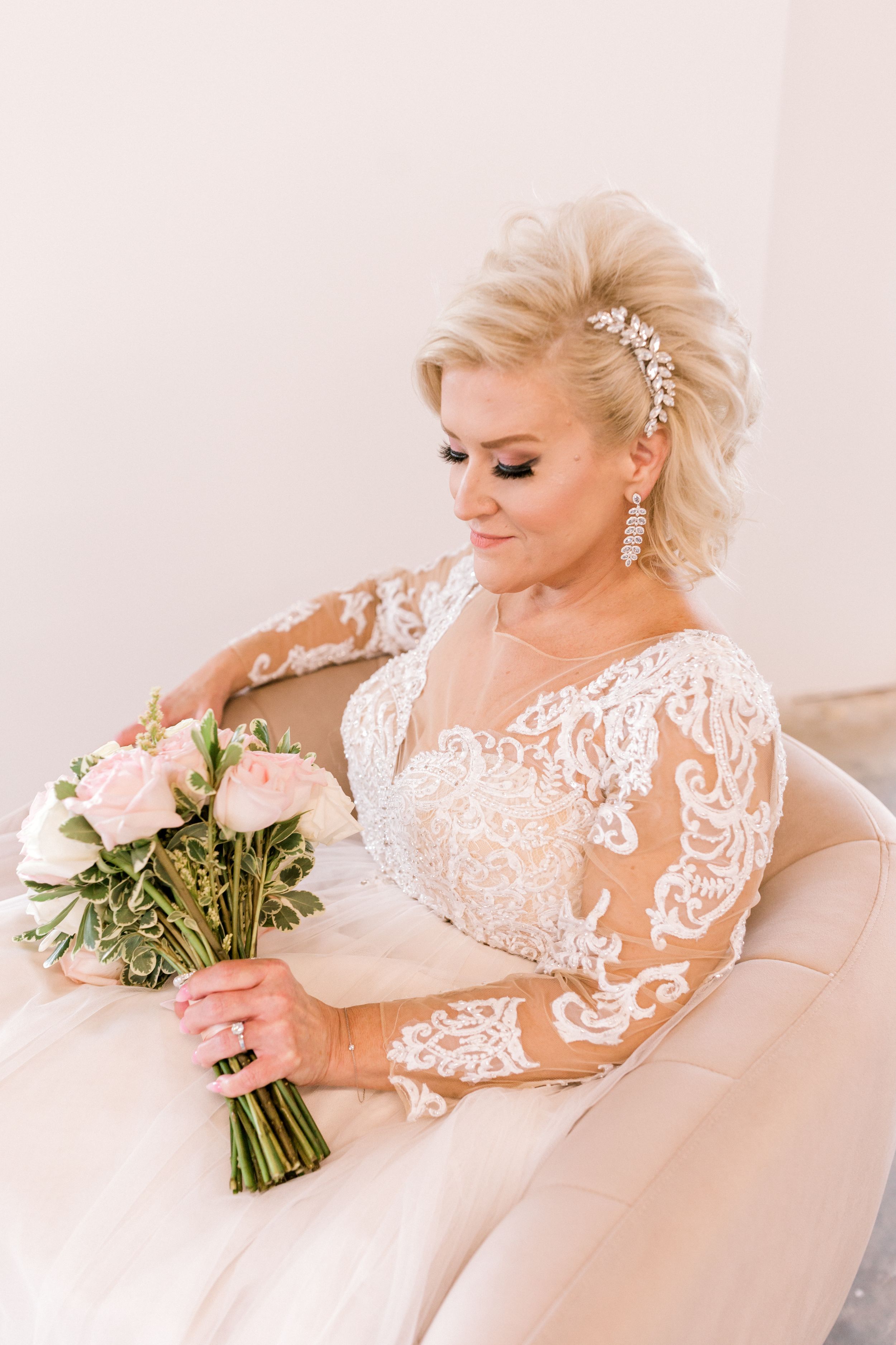 Blonde bride in lace long-sleeve wedding gown holding pink rose bouquet while seated on cream chair.