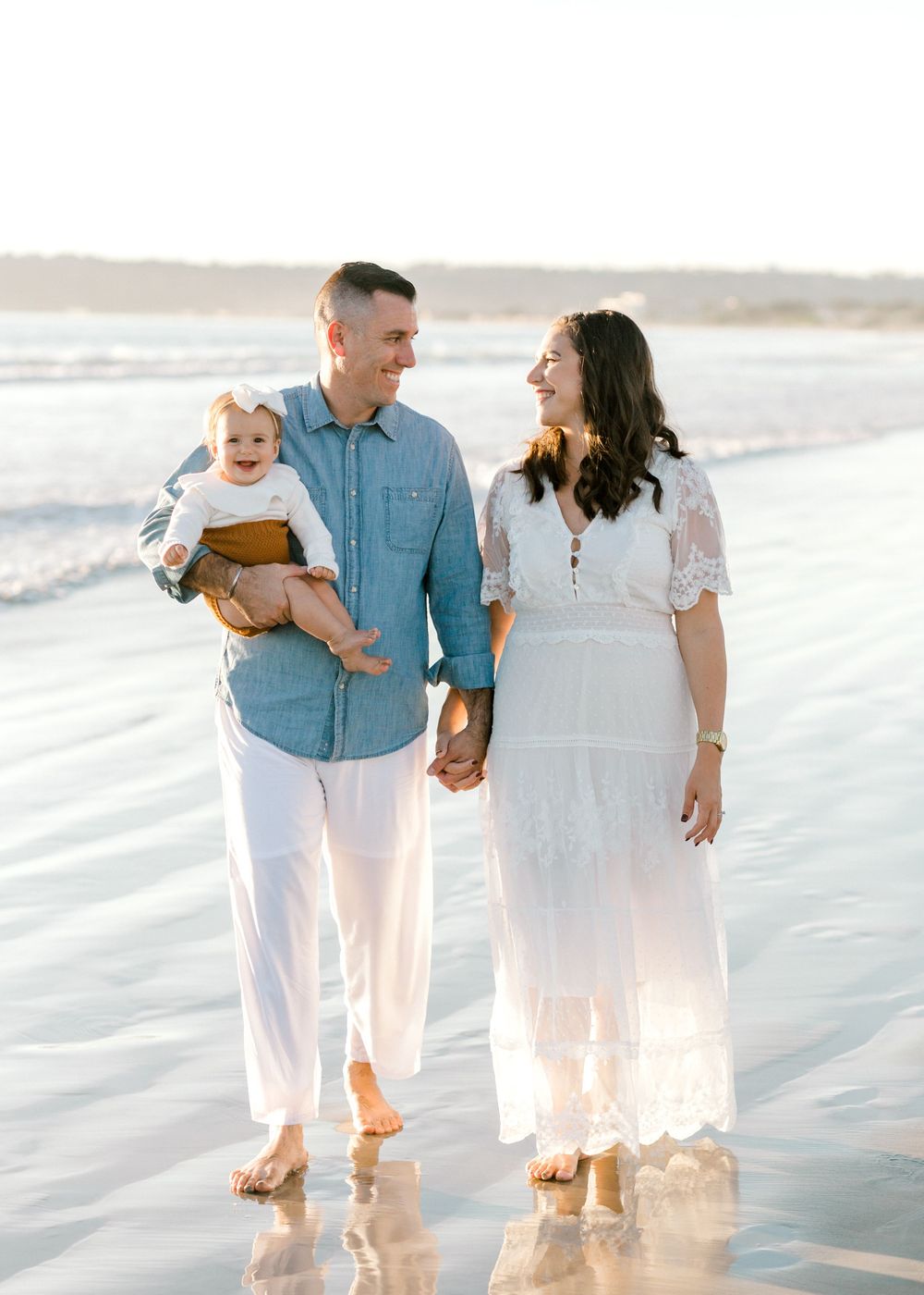 Happy family with baby walking hand-in-hand on reflective beach at golden hour sunset.