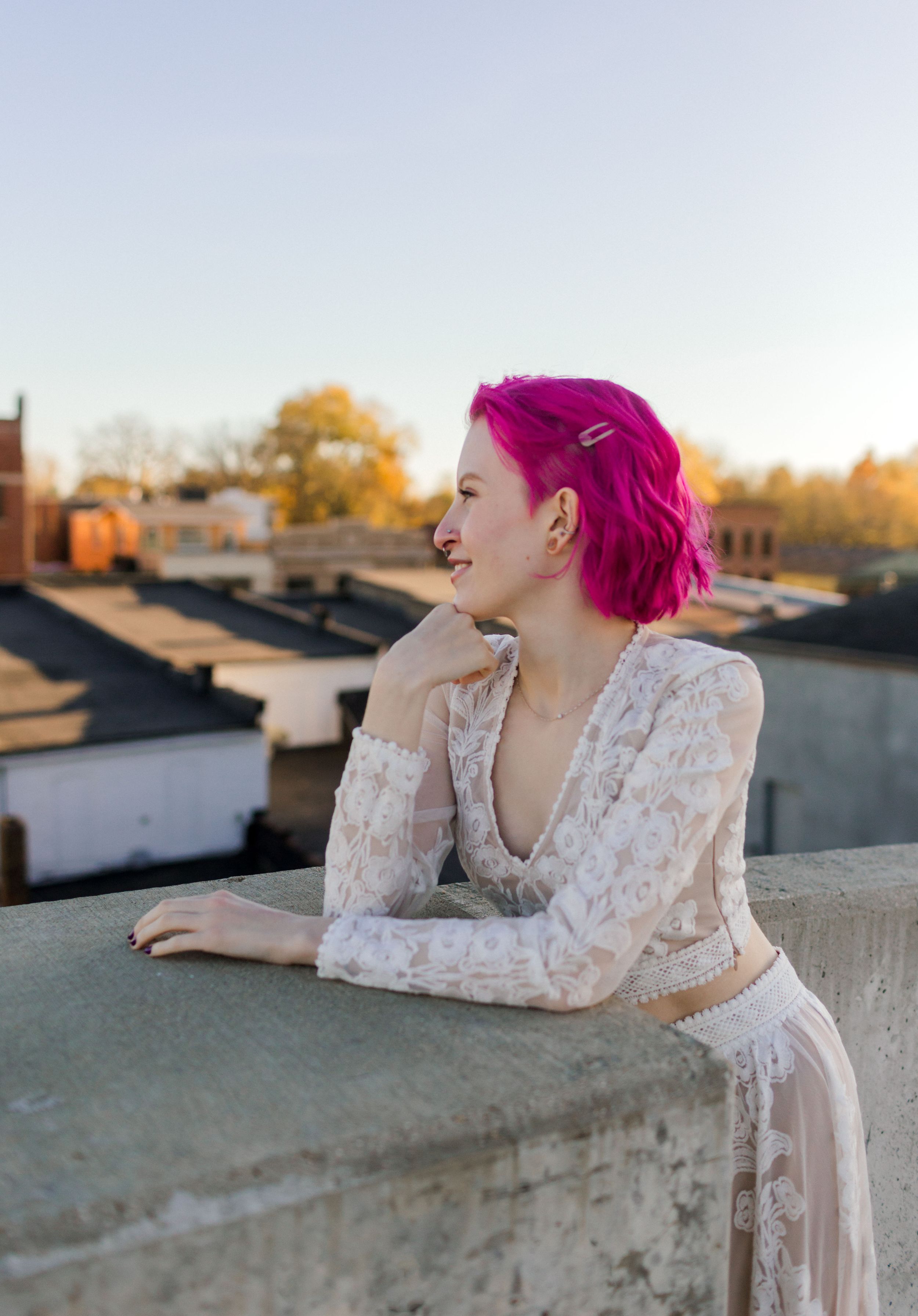 Young woman with vibrant pink hair in white lace outfit leaning on rooftop ledge at sunset.