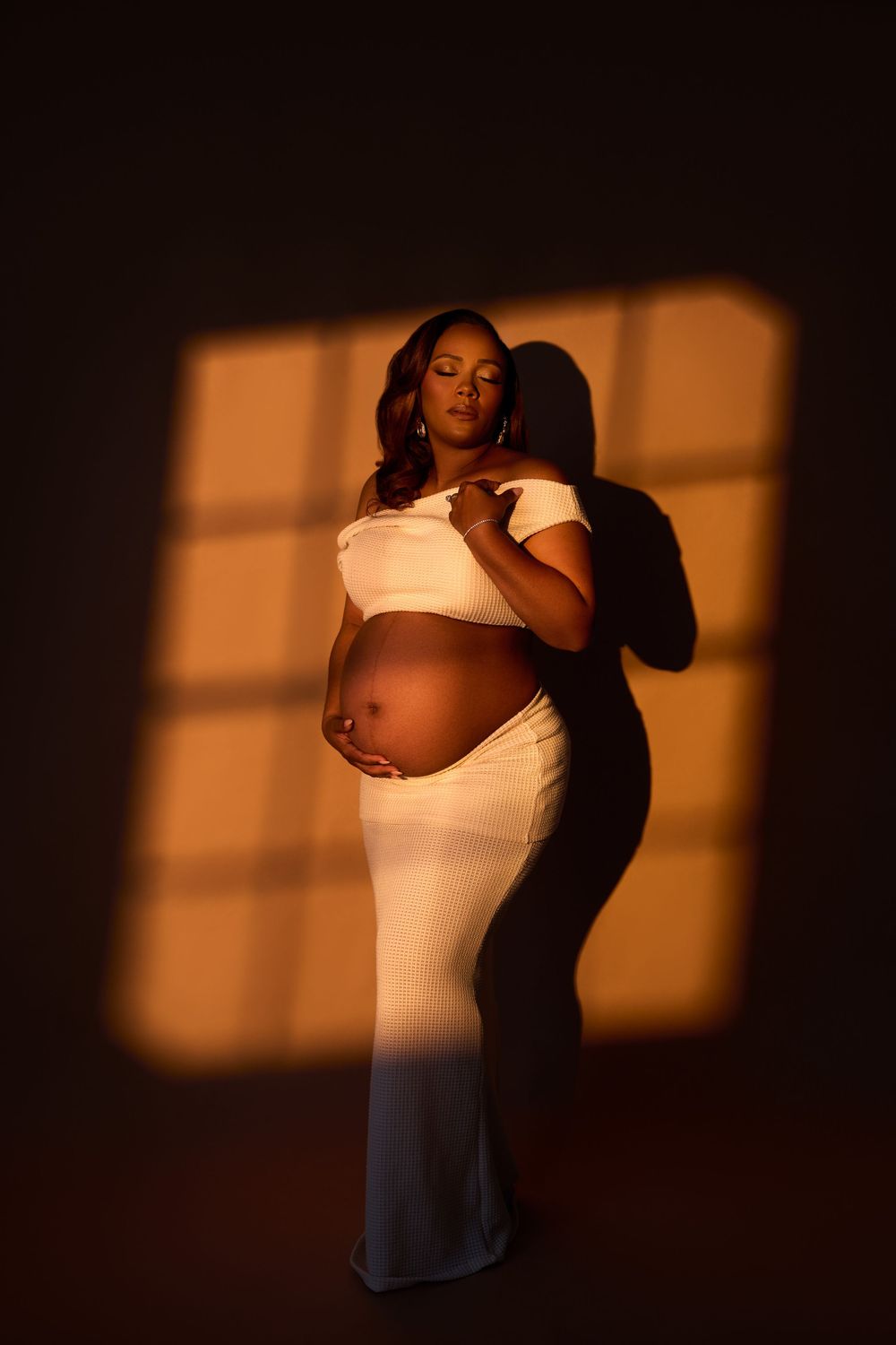 Pregnant Black woman in white crop top and skirt poses gracefully in warm window light against dark background.