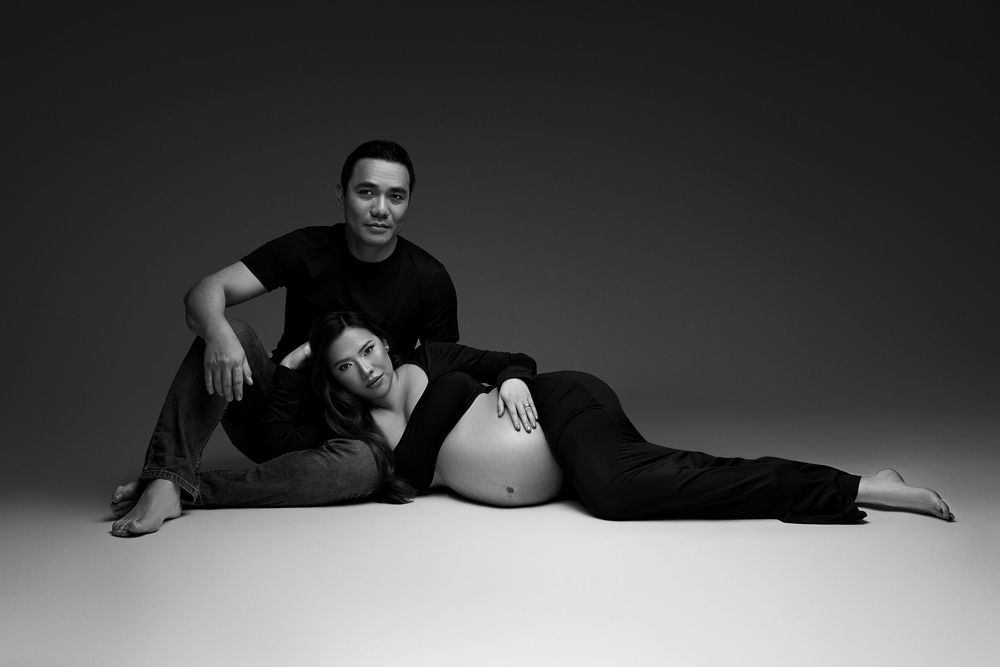 Black and white maternity photo of a couple, woman lying down with baby bump, man sitting beside in a studio. timeless nj maternity photos
