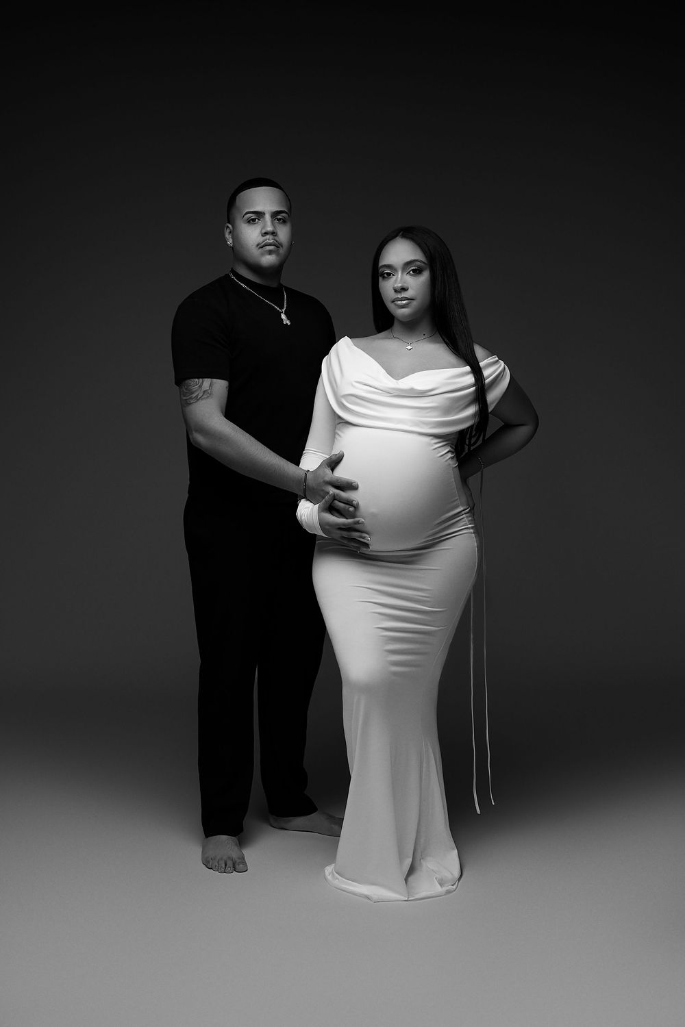 Black and white maternity photo series showing a pregnant woman in a white dress with dramatic studio lighting.