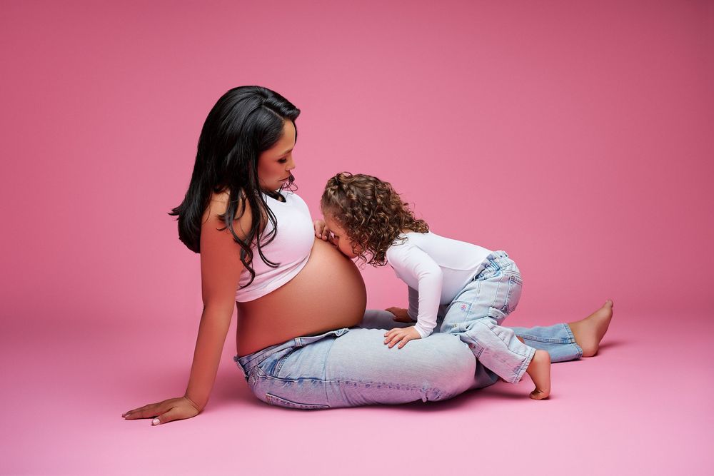 Pregnant woman sitting on pink background with curly-haired child leaning against her baby bump in jeans. Maternity photoshoot ideas