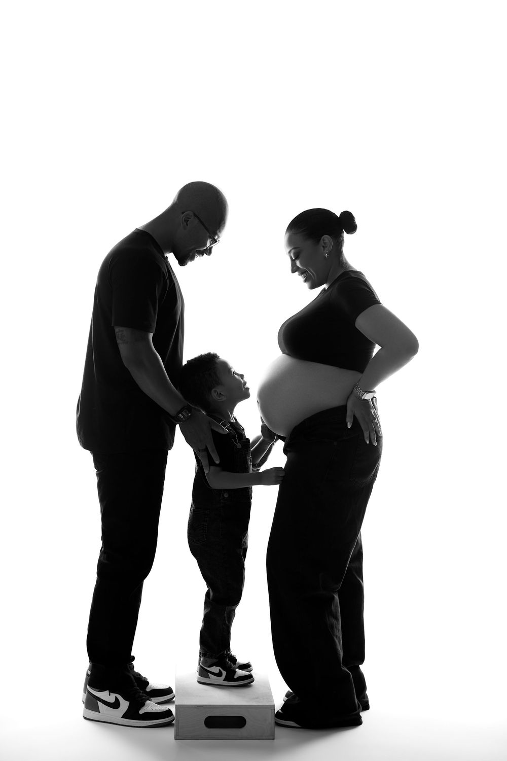 Black and white silhouette of a family with a child standing between parents, one visibly pregnant, against white background.