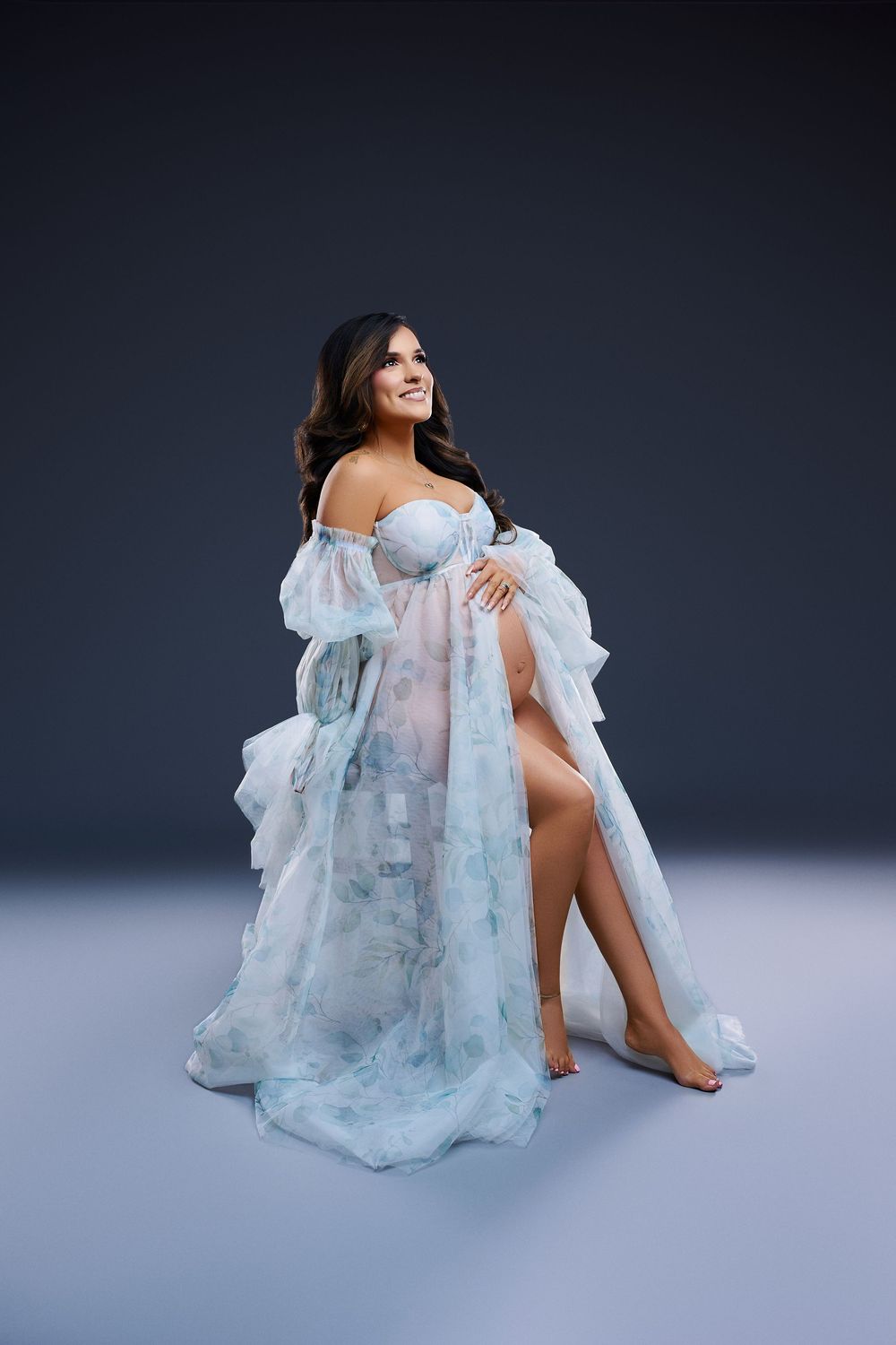Pregnant woman in floral blue sheer maternity gown posing gracefully in studio photography session. Studio Maternity photos