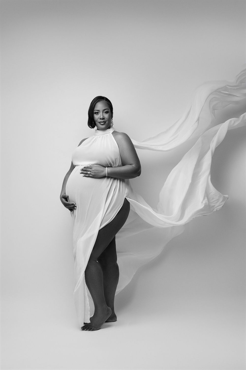 Black and white maternity photo of a pregnant woman in flowing white fabric gown, posing gracefully in a studio.