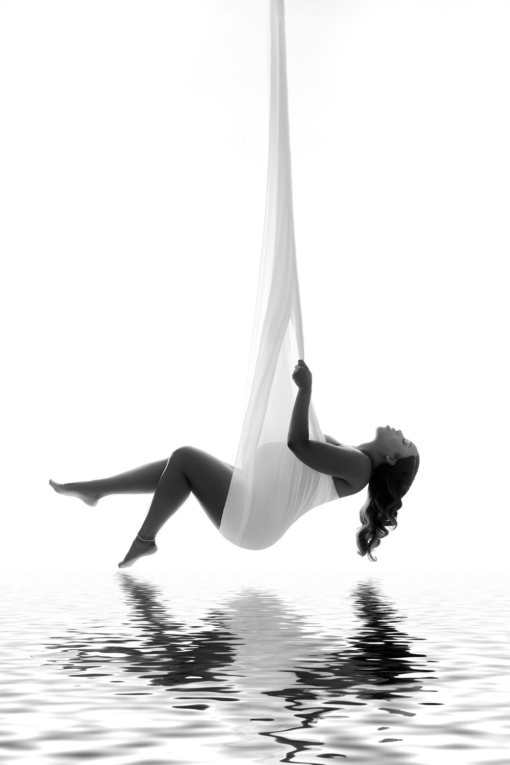 Black and white photo of a woman gracefully suspended in aerial silk, reflected on water below. NJ Maternity Portraits