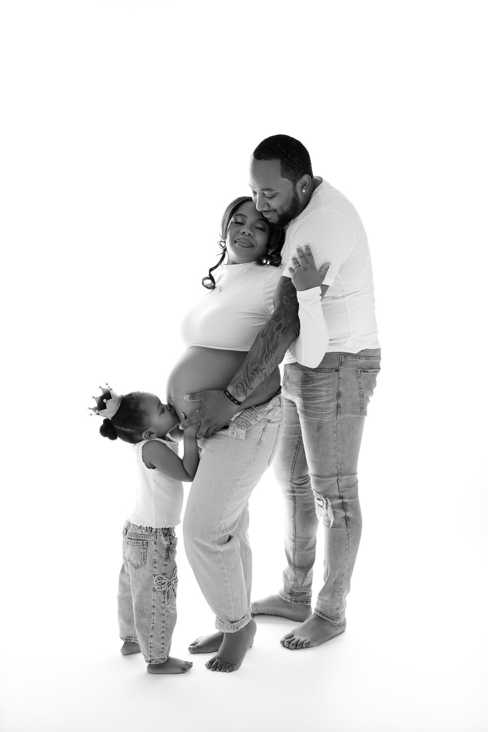 Black and white maternity photo of a couple and toddler, child kissing pregnant belly in a studio setting.