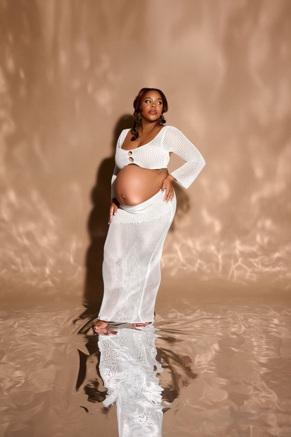 Pregnant woman in white ribbed crop top and sheer maxi skirt posing gracefully in a studio setting.