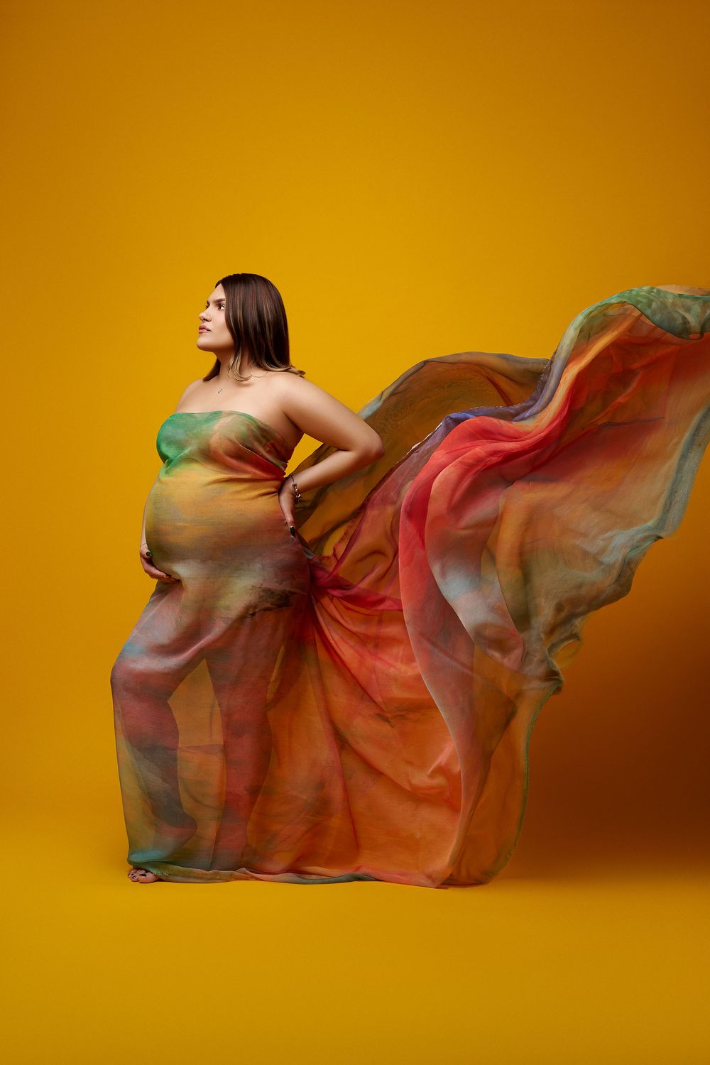 Pregnant woman in flowing colorful chiffon fabric dress posing on yellow background. colorful maternity photos