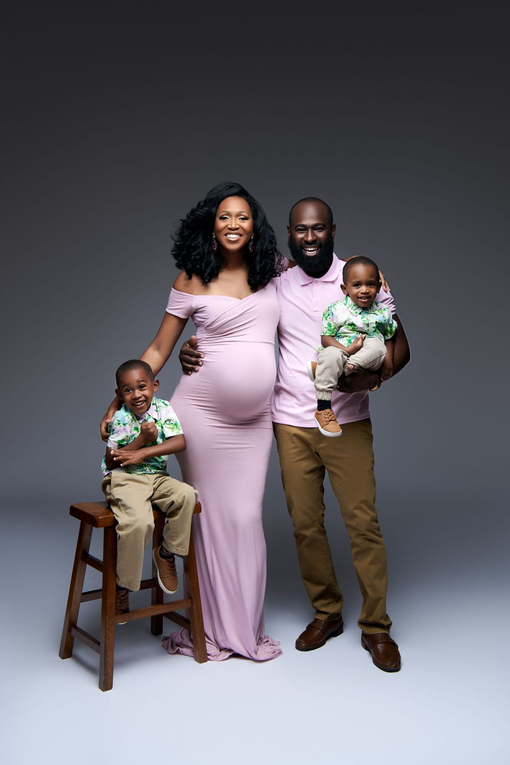 Pregnant woman in pink gown poses with husband and two young sons in matching outfits for a studio family portrait. Maternity studio family