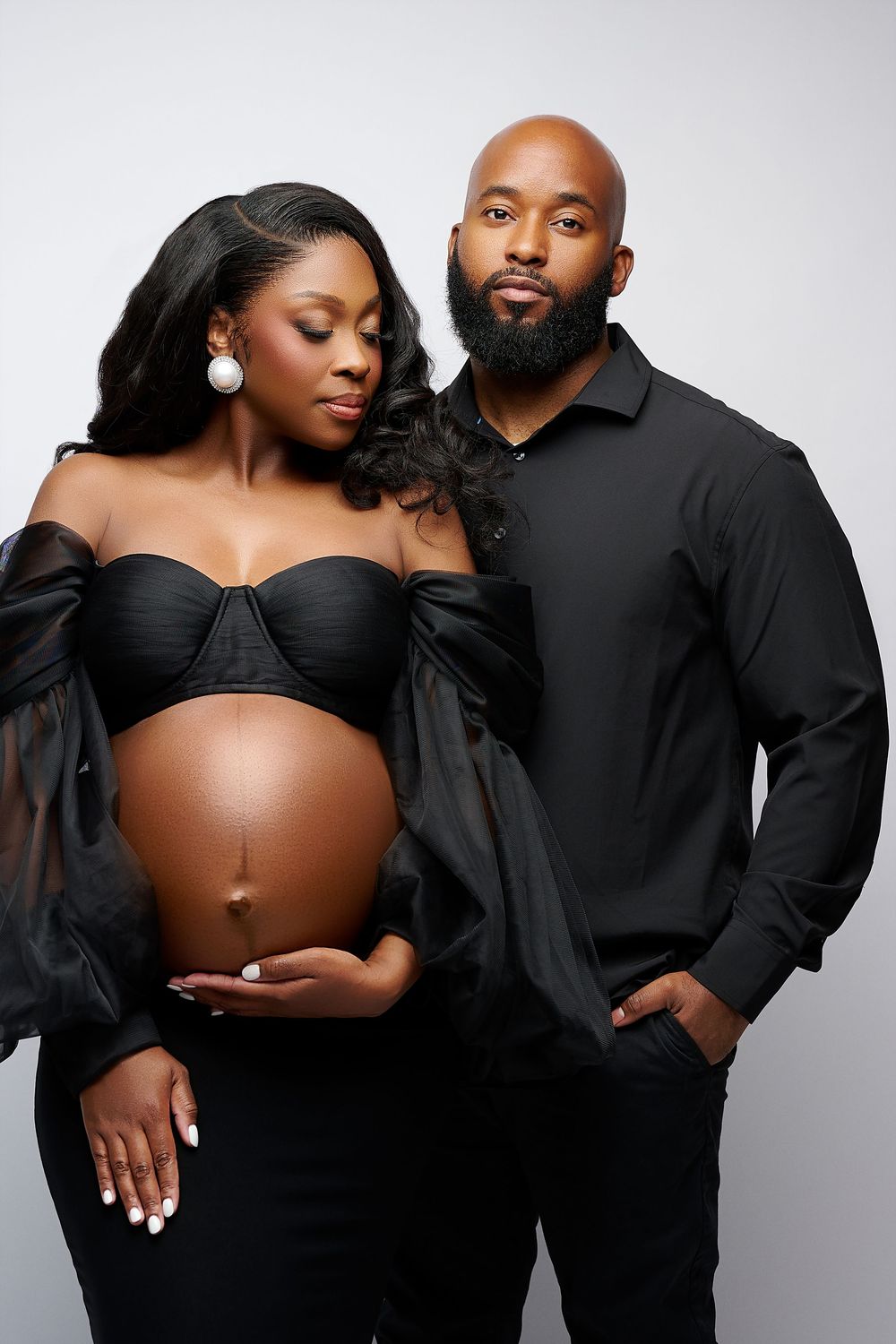 A pregnant Black woman in a black strapless gown poses with her partner in a black shirt for a maternity photo.