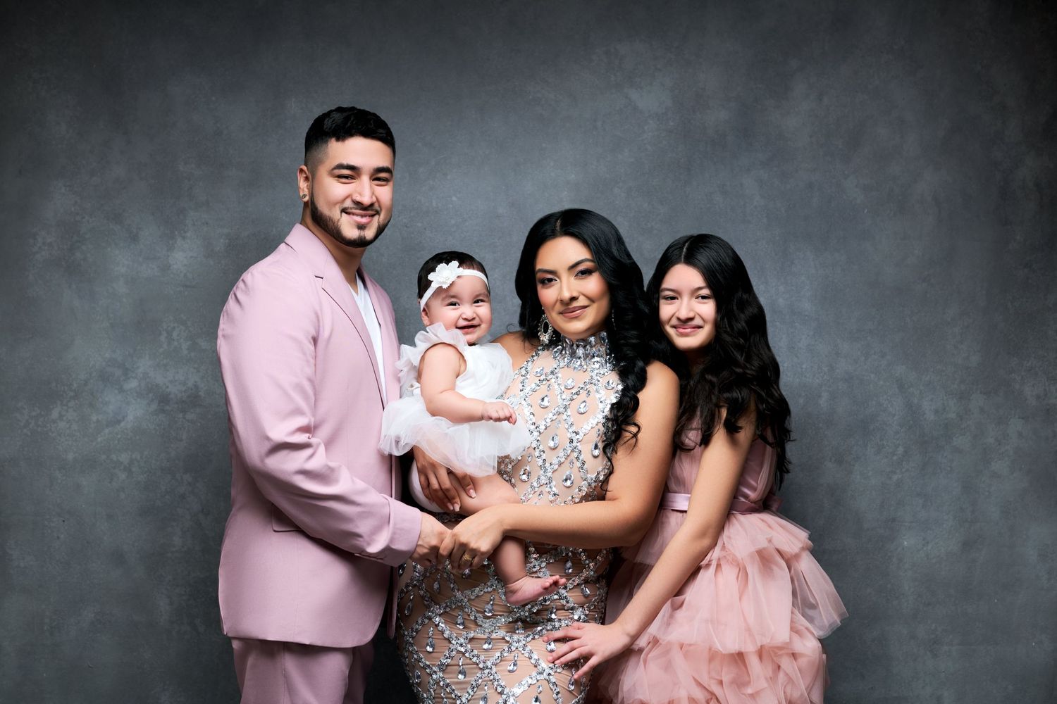 Family poses in coordinating pink outfits for professional studio portraits against gray backdrop. Gricel's Photography NJ