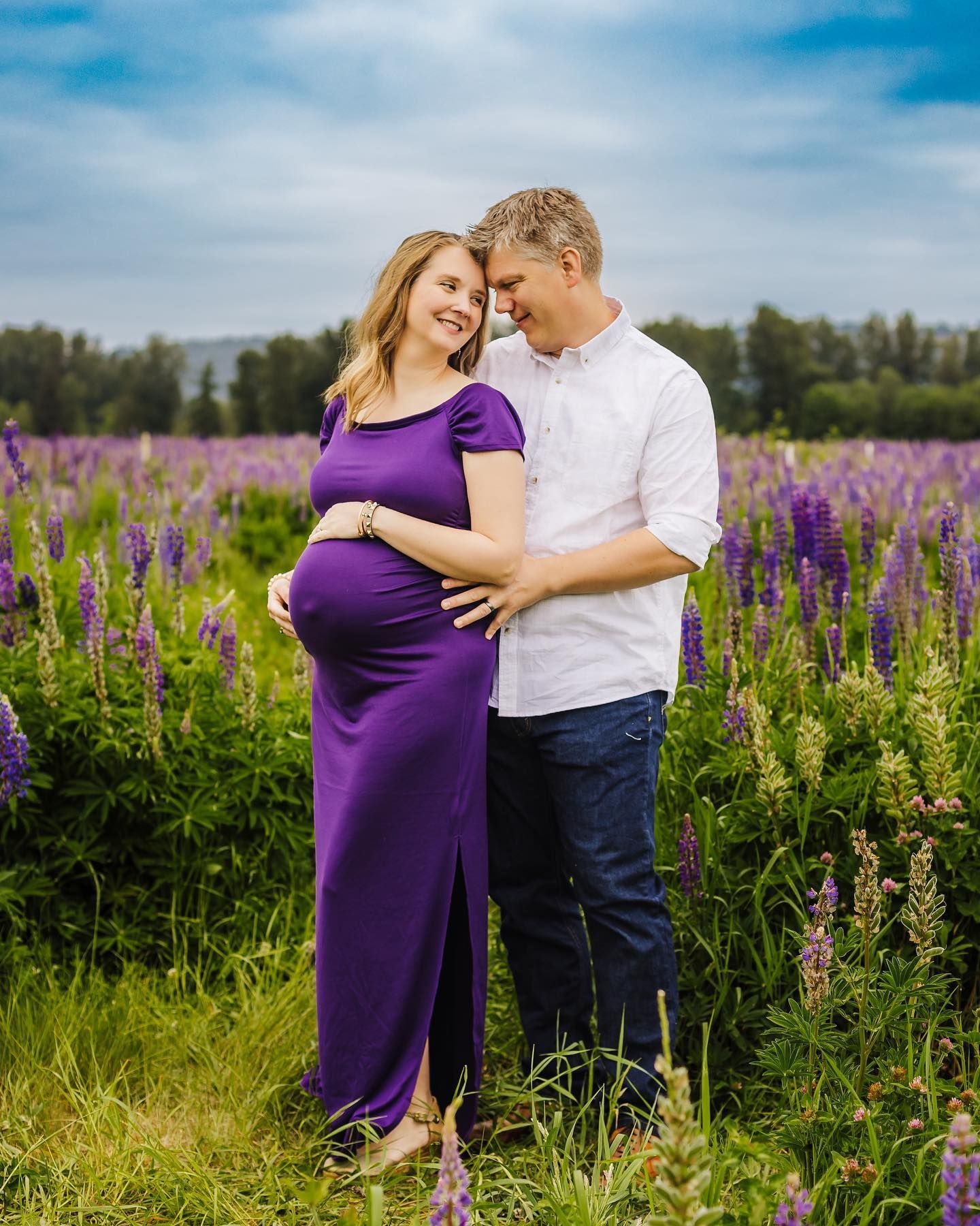 A maternity photo shows a couple embracing in a field of purple wildflowers under blue skies.