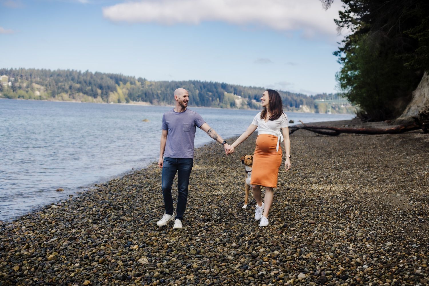 A couple walks hand in hand along a rocky beach shore with water and forested hills in the background.