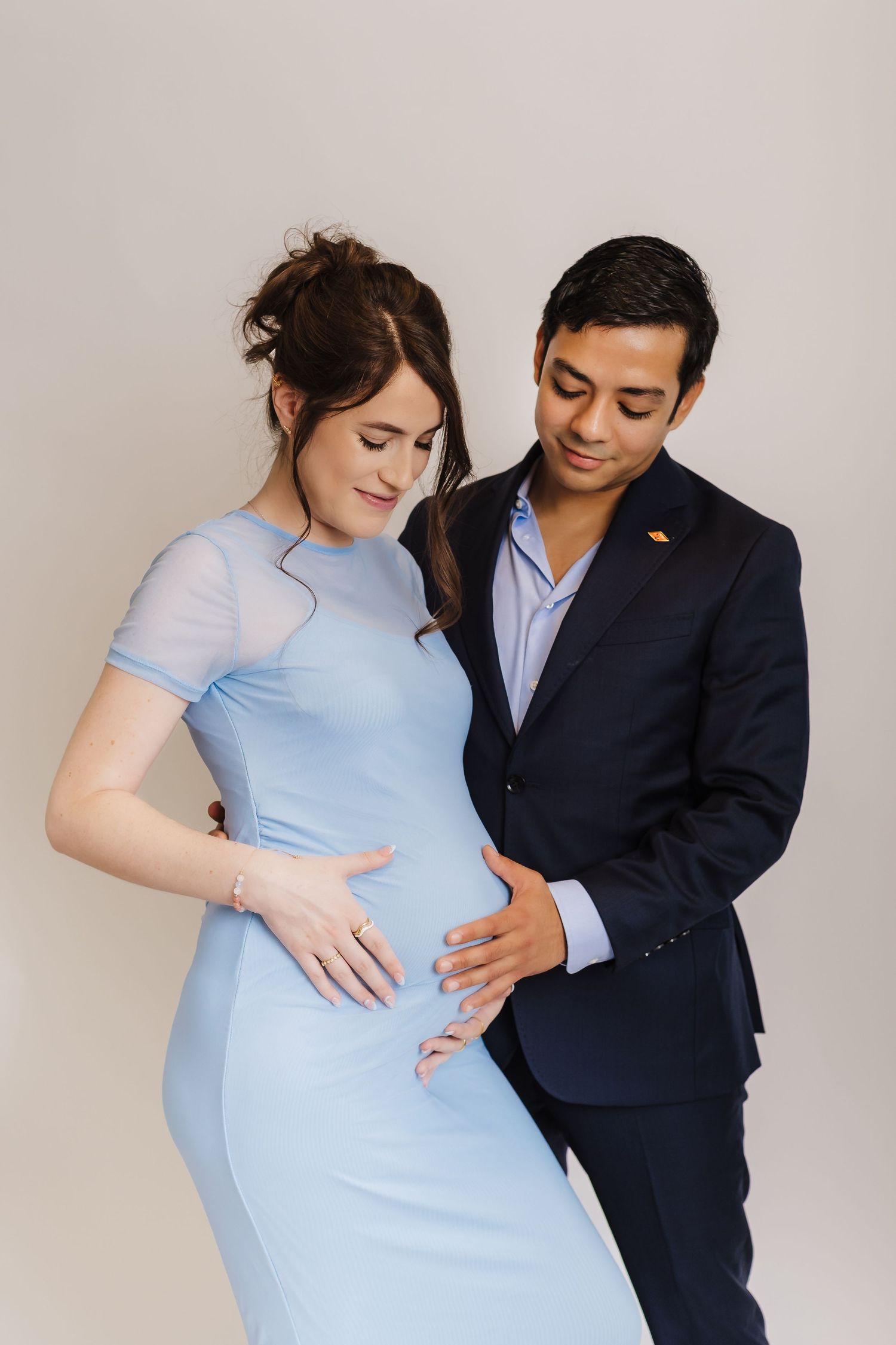 Couple in blue dress and navy suit embrace while sharing a tender pregnancy moment against a neutral background.