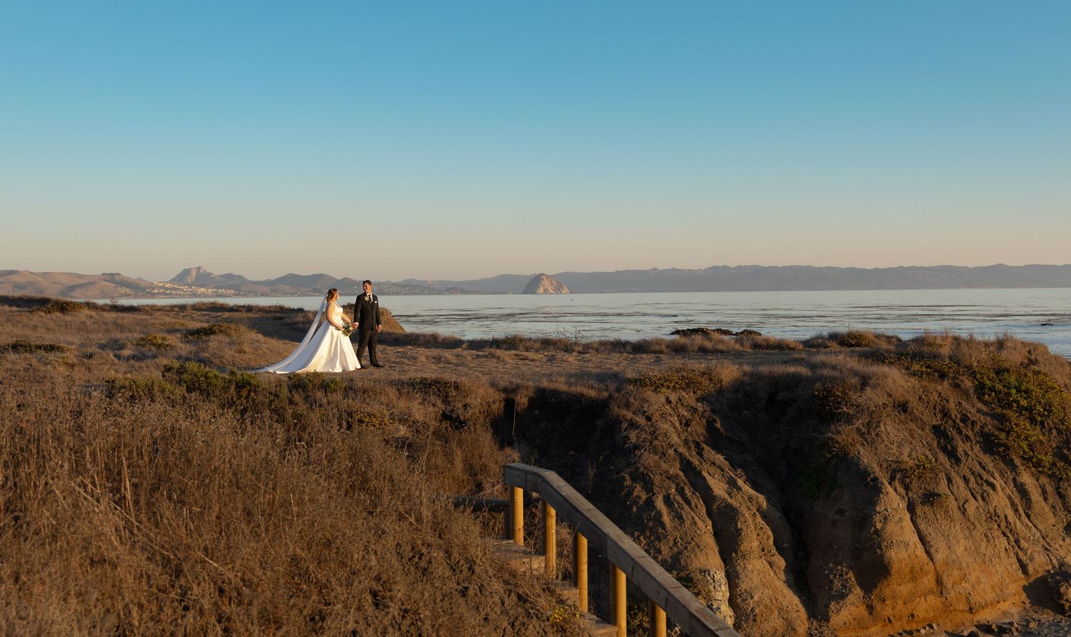 Bride and groom walk hand and hand in Cayucos, California on the cliffside overlooking ocean views.