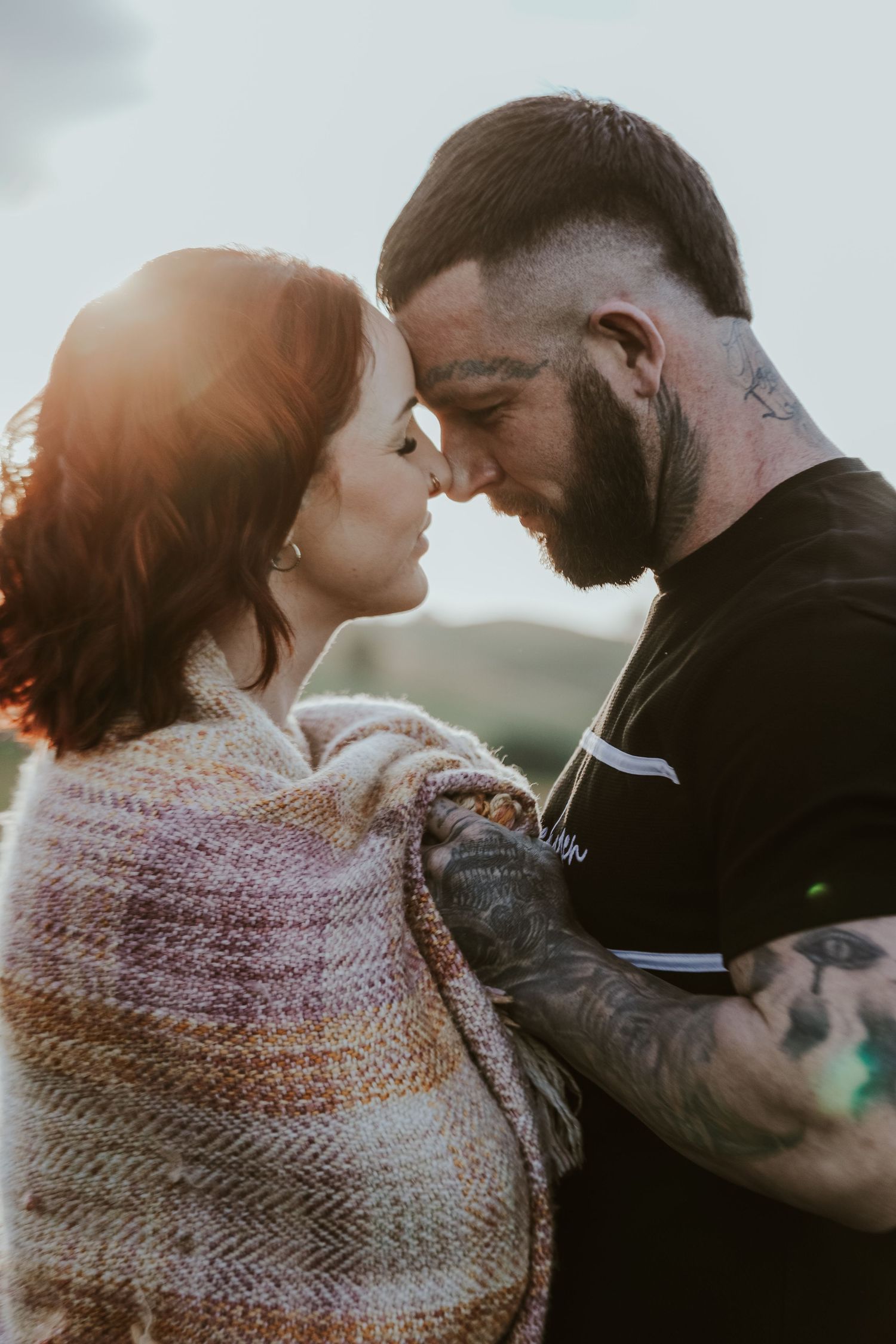 Romantic couple embraces in outdoor photoshoot wearing cozy knit scarf at sunset.