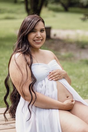 A maternity portrait in a white flowing dress against a lush green background.
