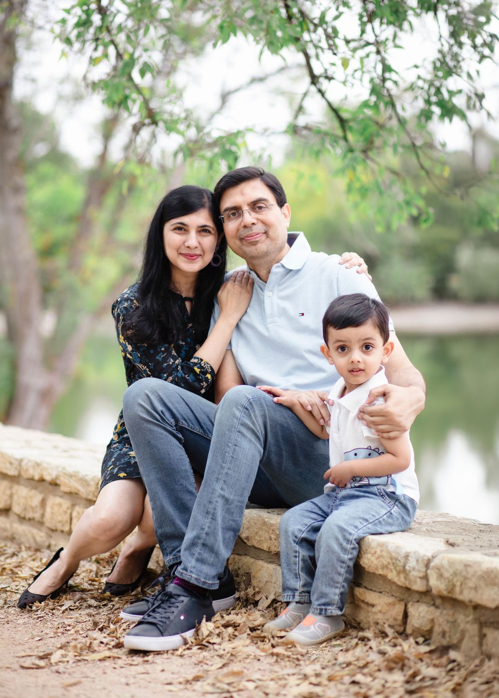 A family sitting together on a stone wall near a pond surrounded by green trees in a tranquil outdoor setting. Well branch Fall Family Mini