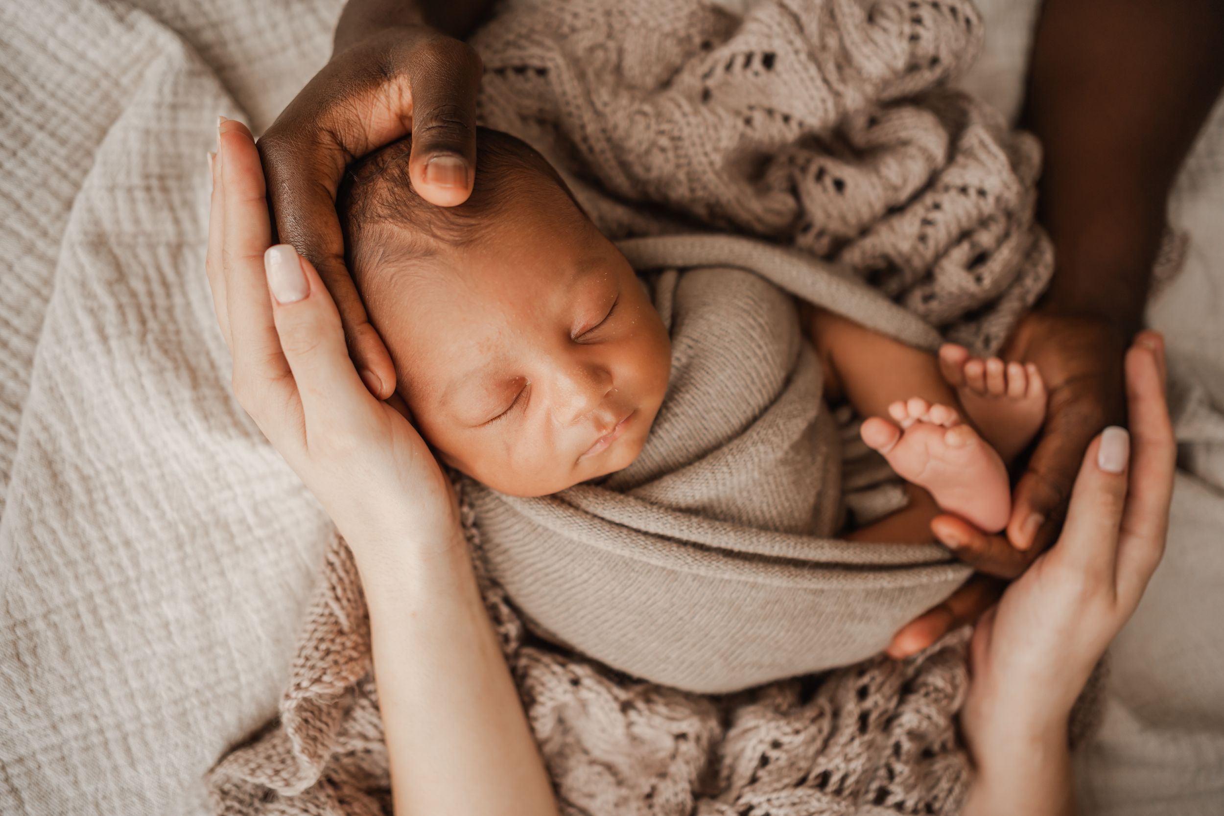 Newborn baby swaddled in neutral tones, cradled by both parents' hands during an in-home family photography session in Portland, Dorset.