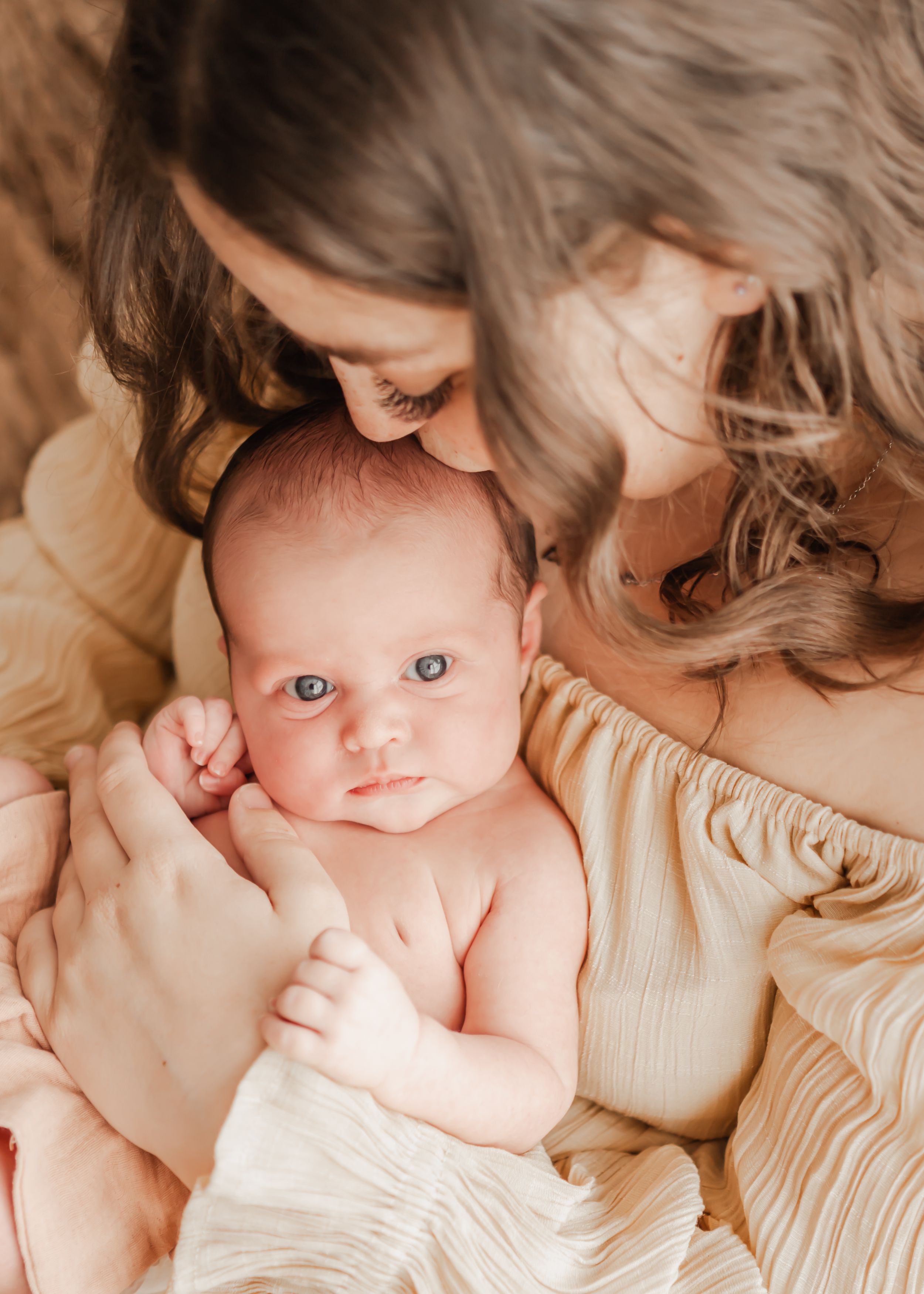 Mother kissing her newborn baby's head as the baby gazes directly at the photographer, during an in-home newborn session in Portland, Dorset