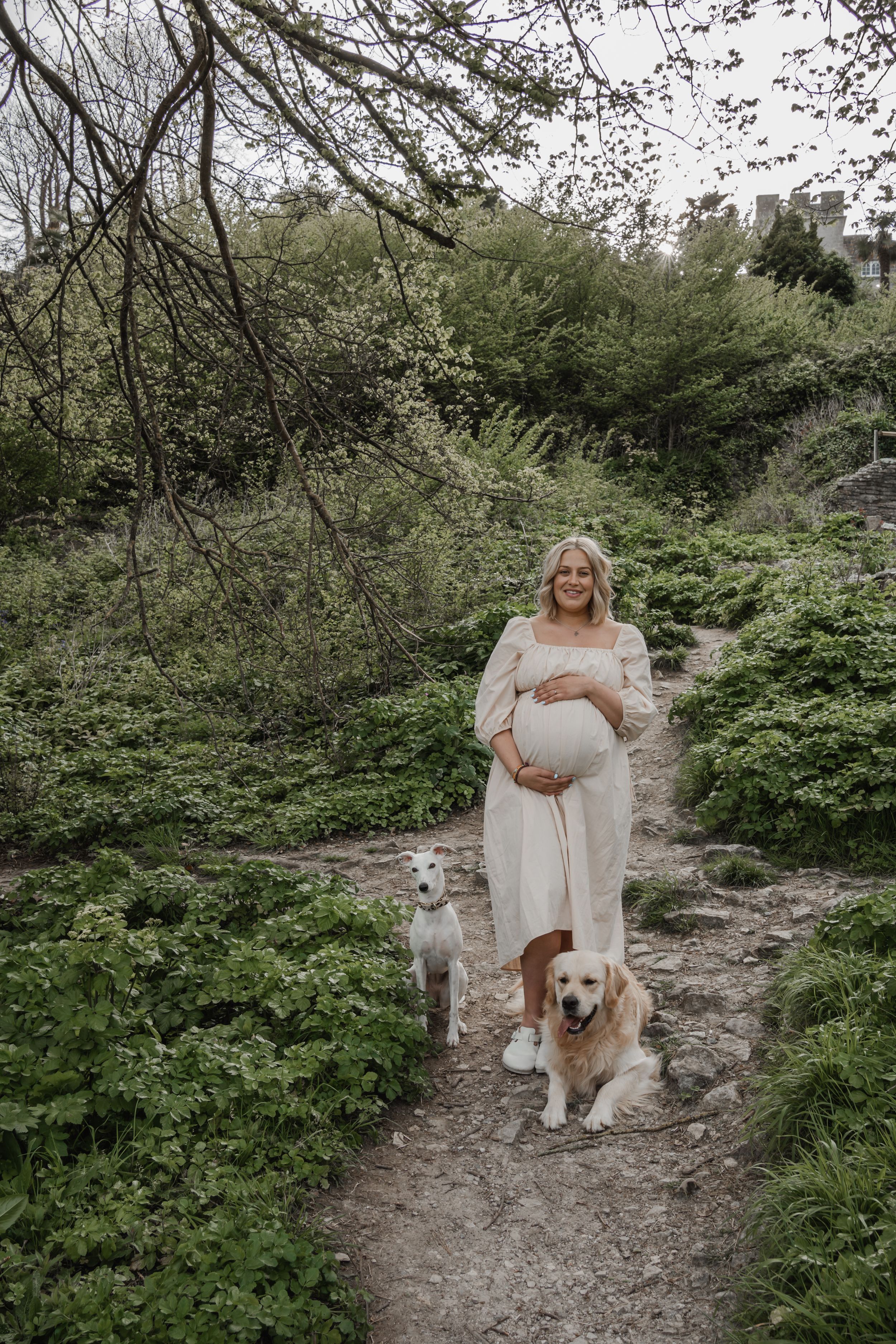 Image taken by an in-home newborn photographer of a Mum-to-be in a cream dress walking her two dogs on a forest trail in Portland, Dorset.