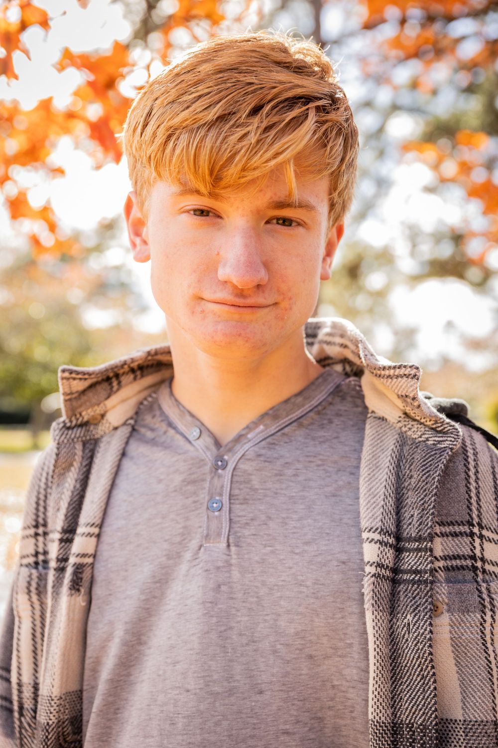 A person wearing a grey henley shirt and plaid flannel jacket poses outdoors with autumn foliage in the background.