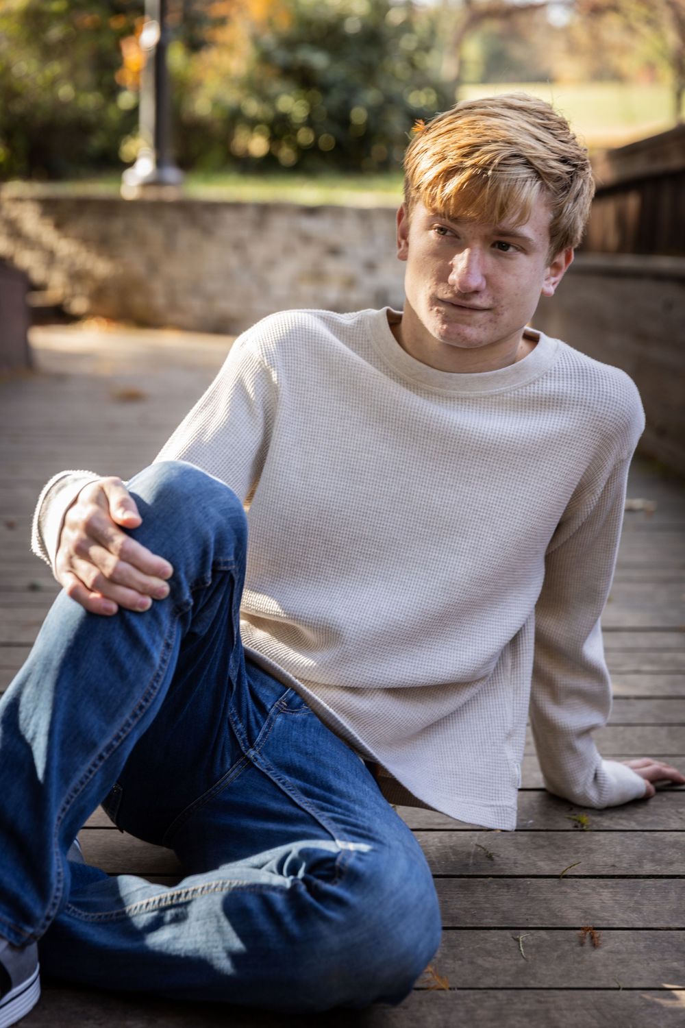 Person in blue jeans and cream sweater sits casually on wooden deck during autumn afternoon.