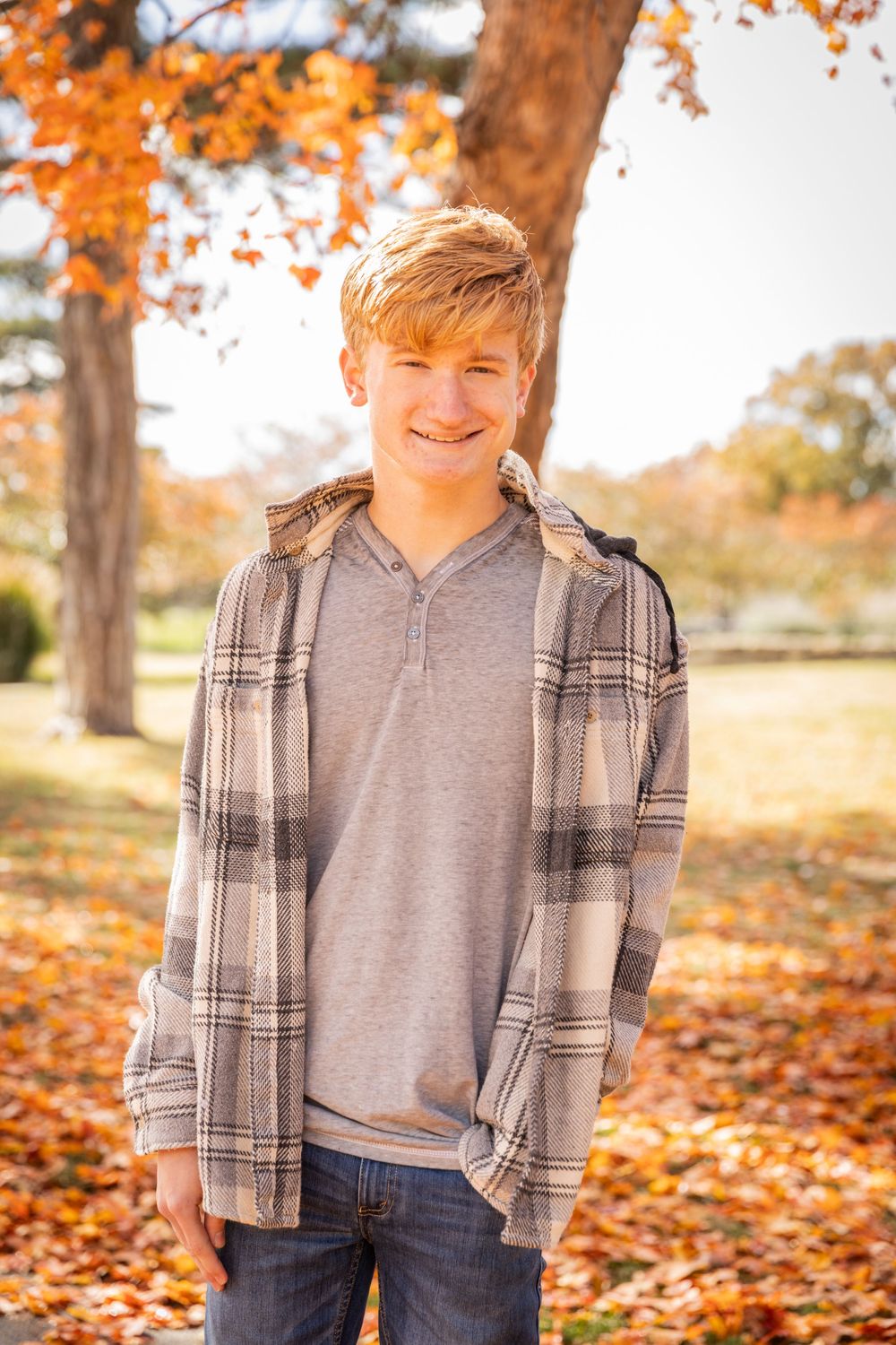 Young person in plaid flannel shirt and gray t-shirt poses outdoors among vibrant orange autumn leaves.