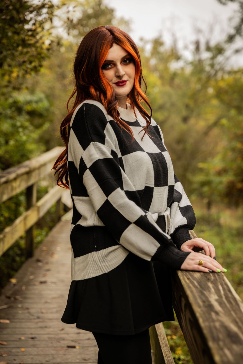 Person in black and white checkered sweater posing on wooden bridge in fall setting.