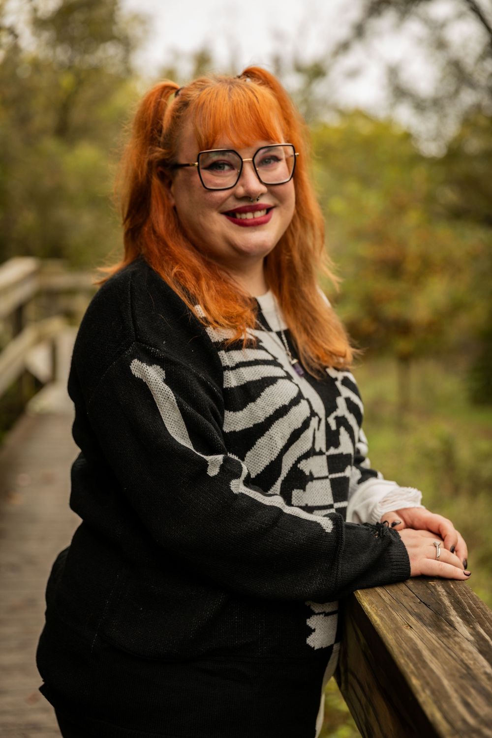 Person wearing skeleton print sweater and glasses poses on wooden bridge in autumn setting.
