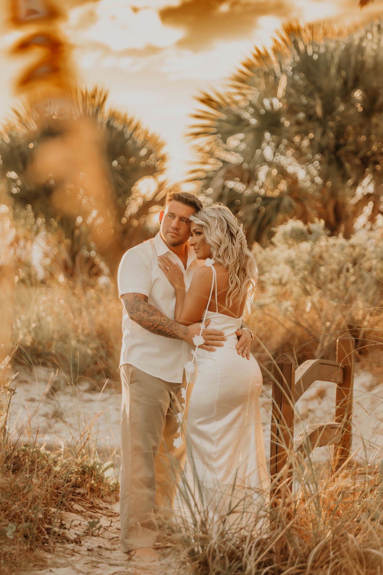 Intimate beach wedding portrait of couple embracing at golden hour among palm trees.