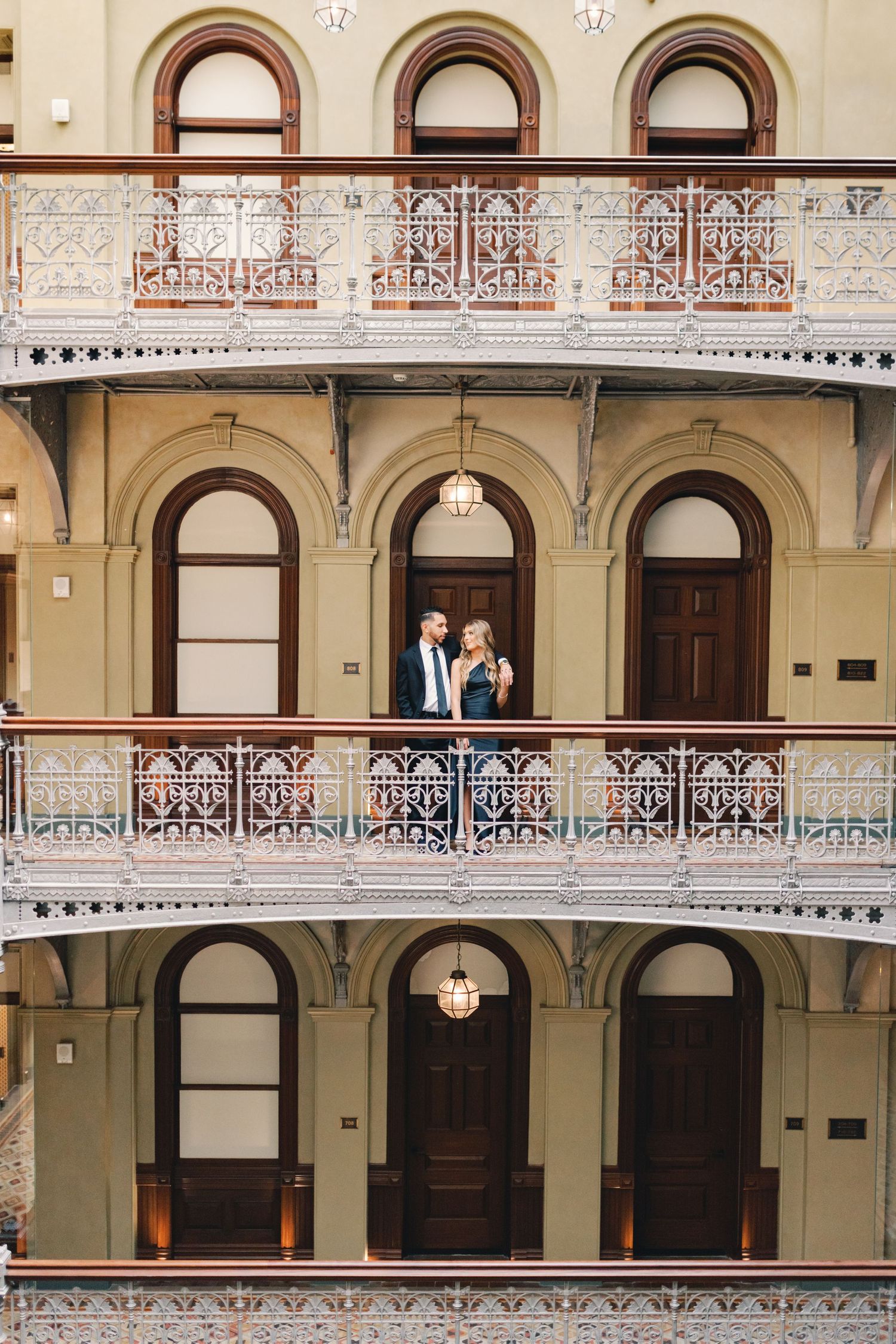 Romantic couple standing on an ornate Victorian-style balcony of a historic multi-story building with arched doorways.