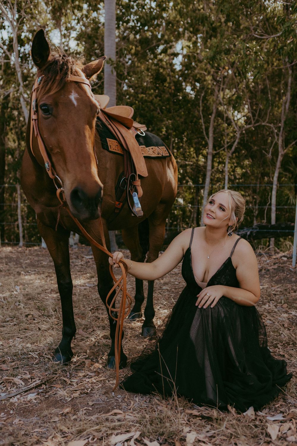 Woman in black gown kneeling beside saddled chestnut horse in Australian bushland setting.